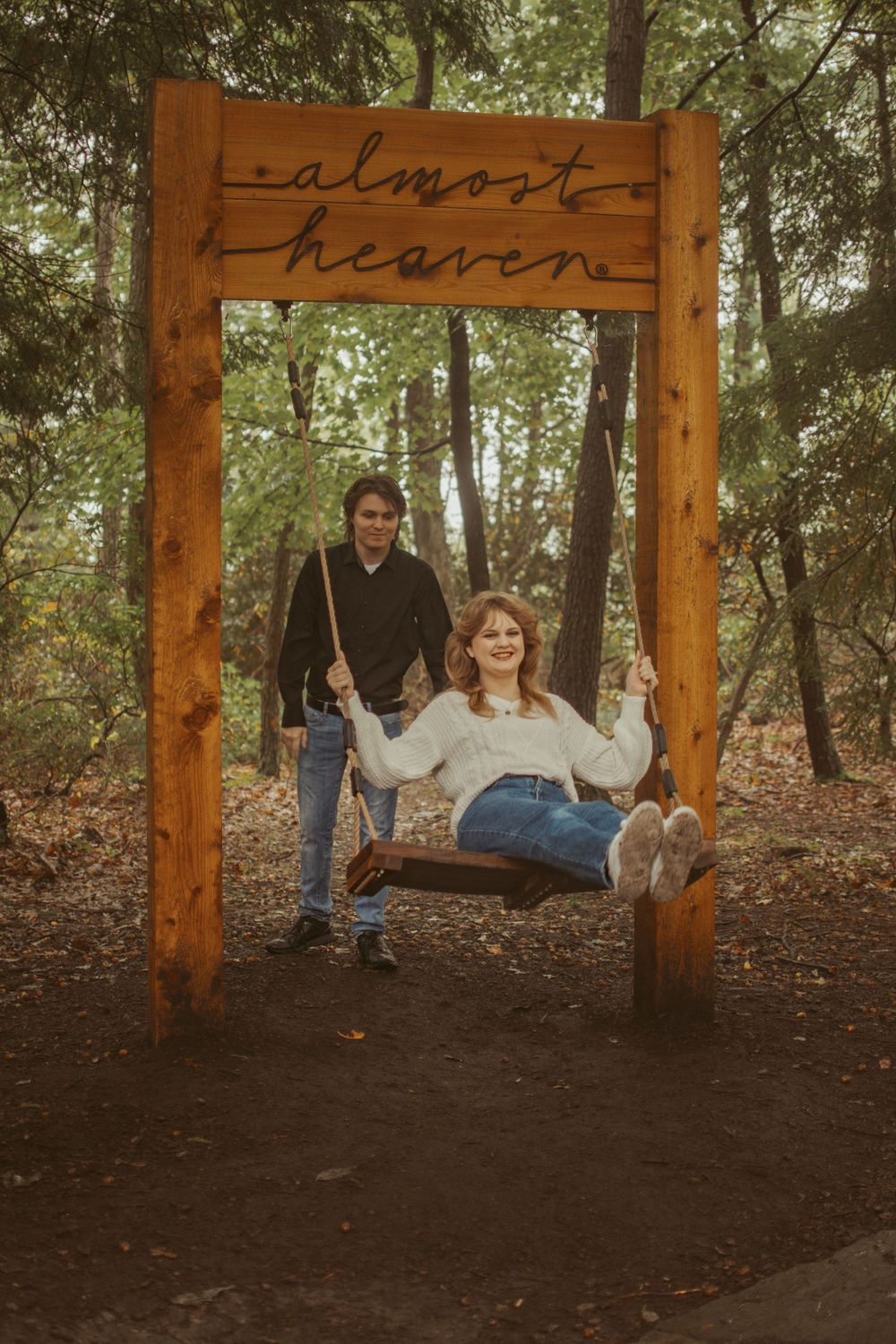 A rustic wooden swing frame with 'almost heaven' text displays a couple in casual attire enjoying an autumn forest setting.