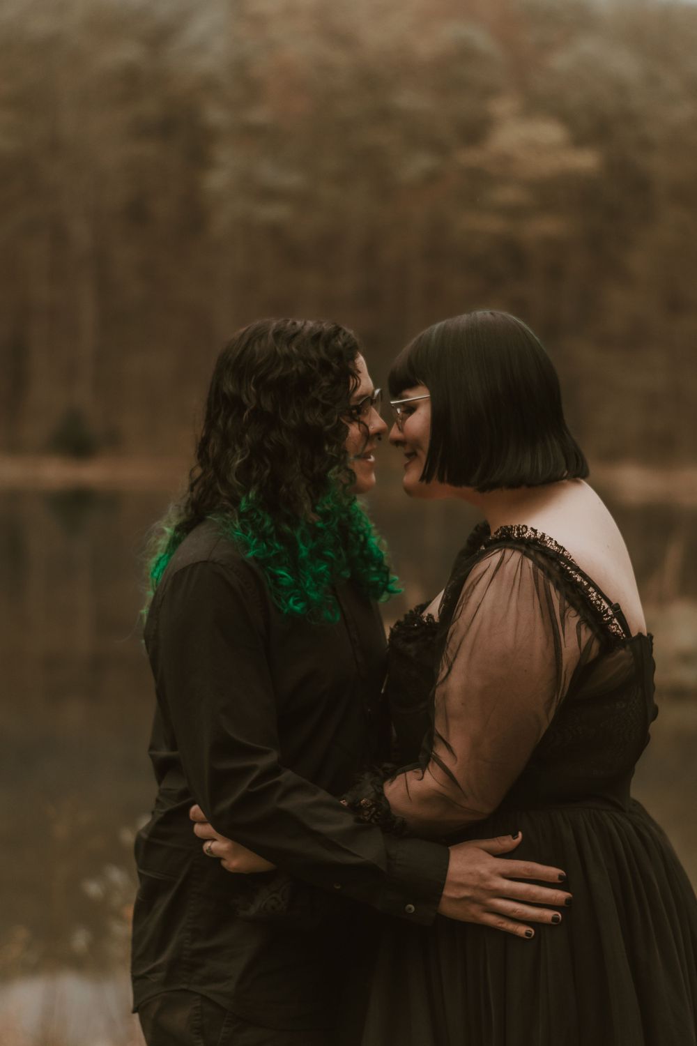 Romantic moment between couple in gothic attire against moody lake backdrop.