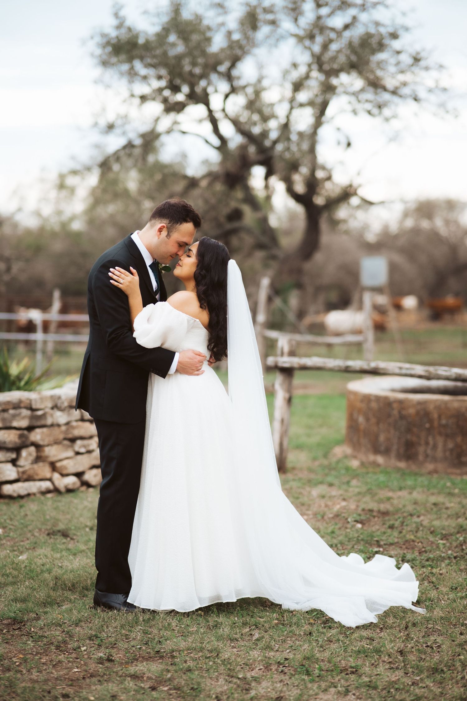 A romantic wedding portrait captures a couple embracing near a rustic stone wall and tree.