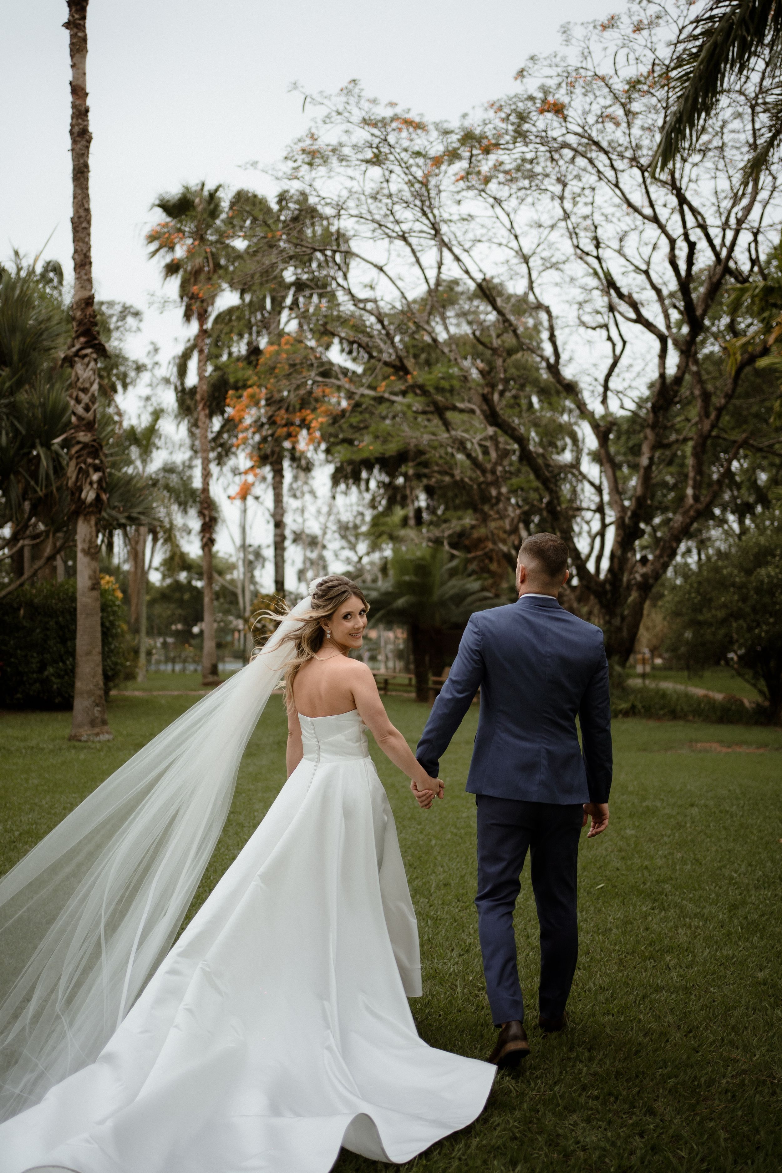 Couple walking hand in hand through tropical garden with bride's flowing veil trailing behind.