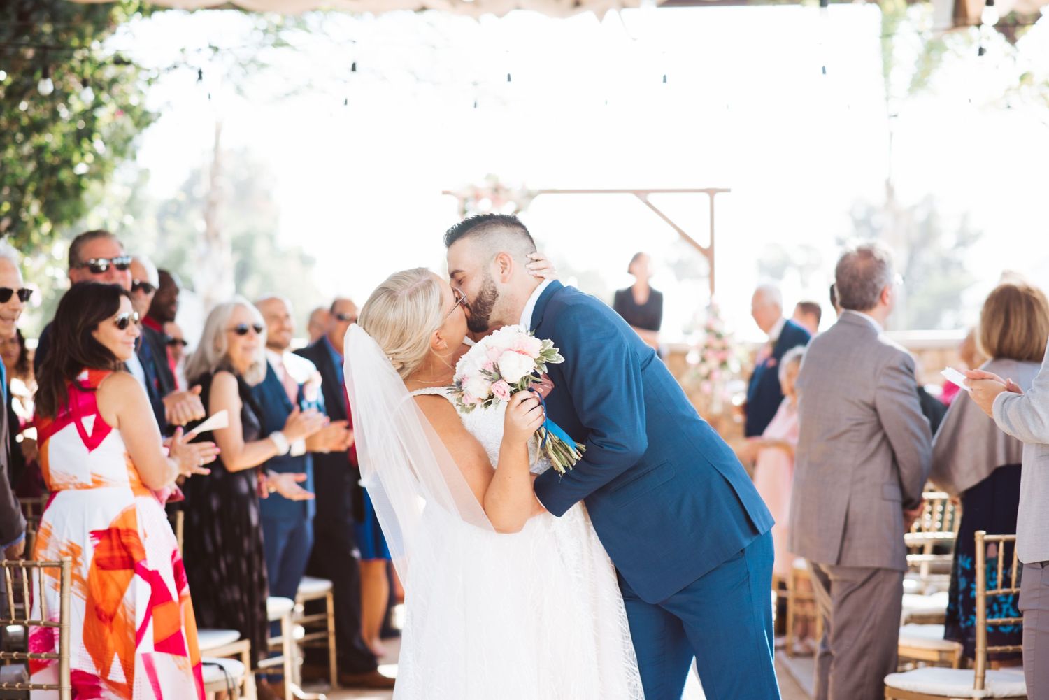 Newlyweds share their first kiss at outdoor wedding ceremony surrounded by guests.