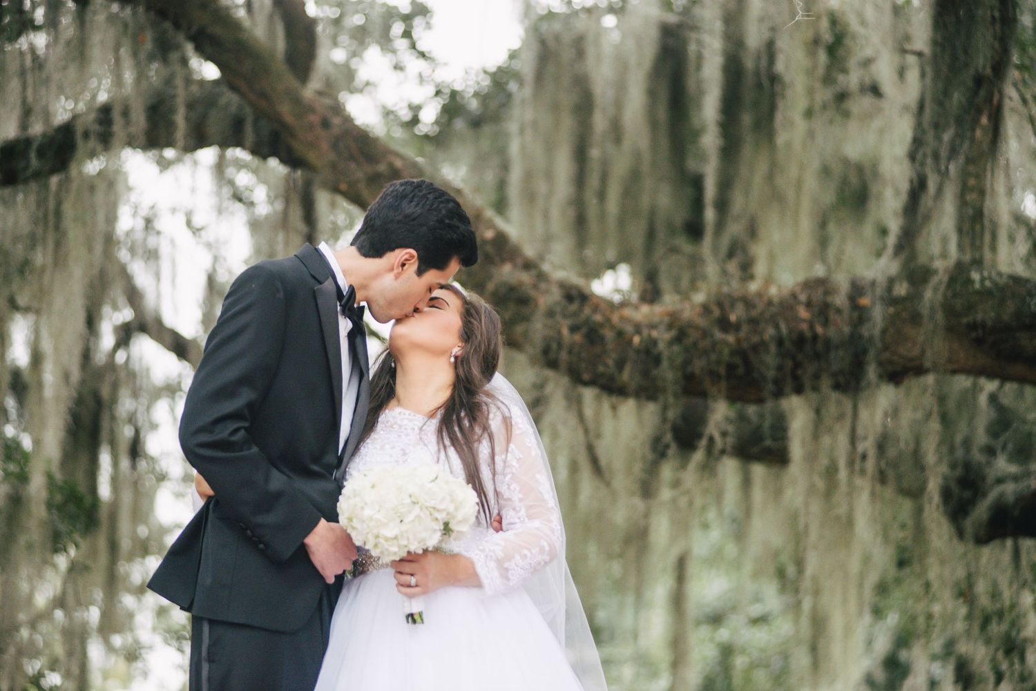 A romantic wedding kiss under Spanish moss in a Southern garden setting.