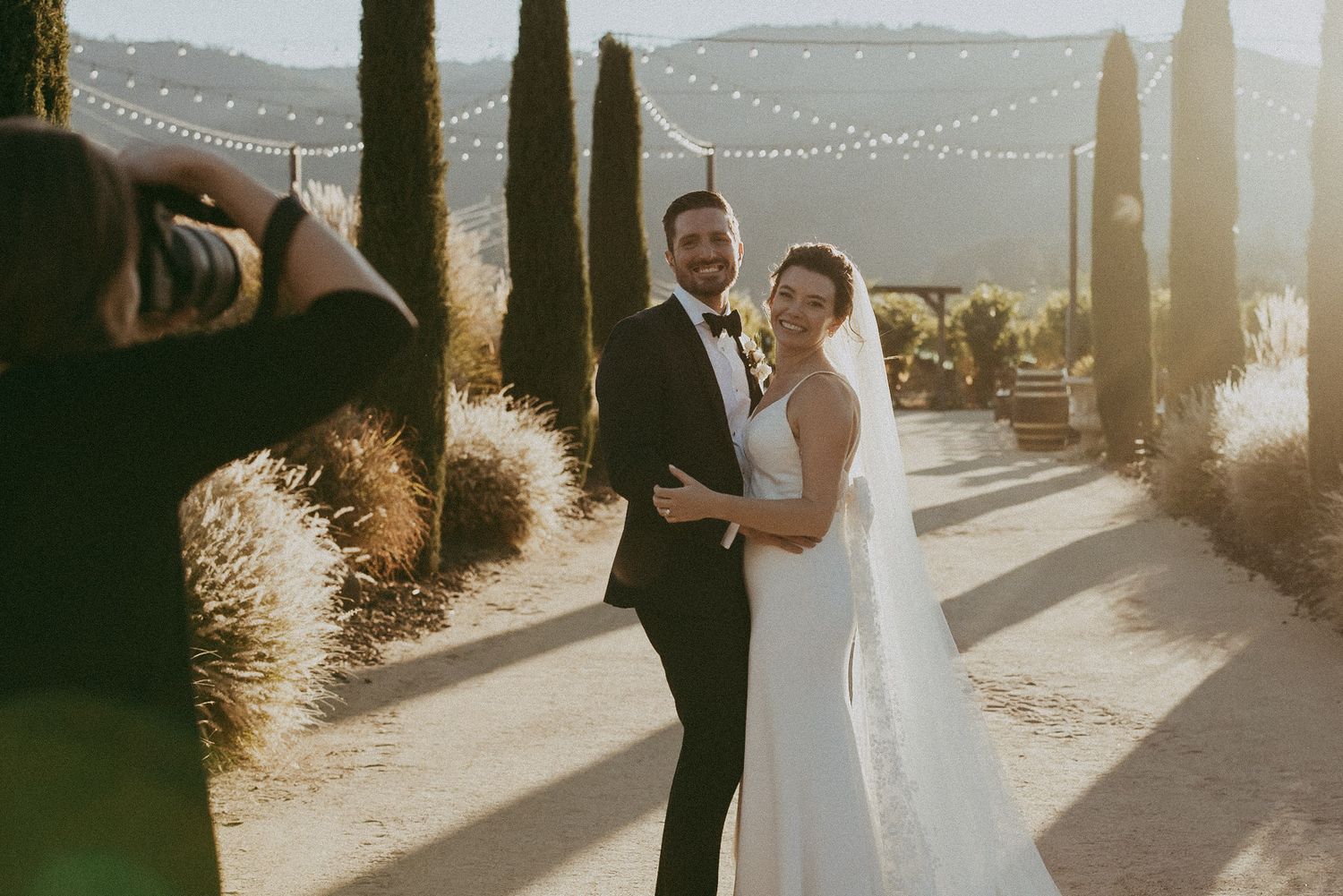 Golden hour wedding portrait against a scenic hilltop backdrop with string lights.