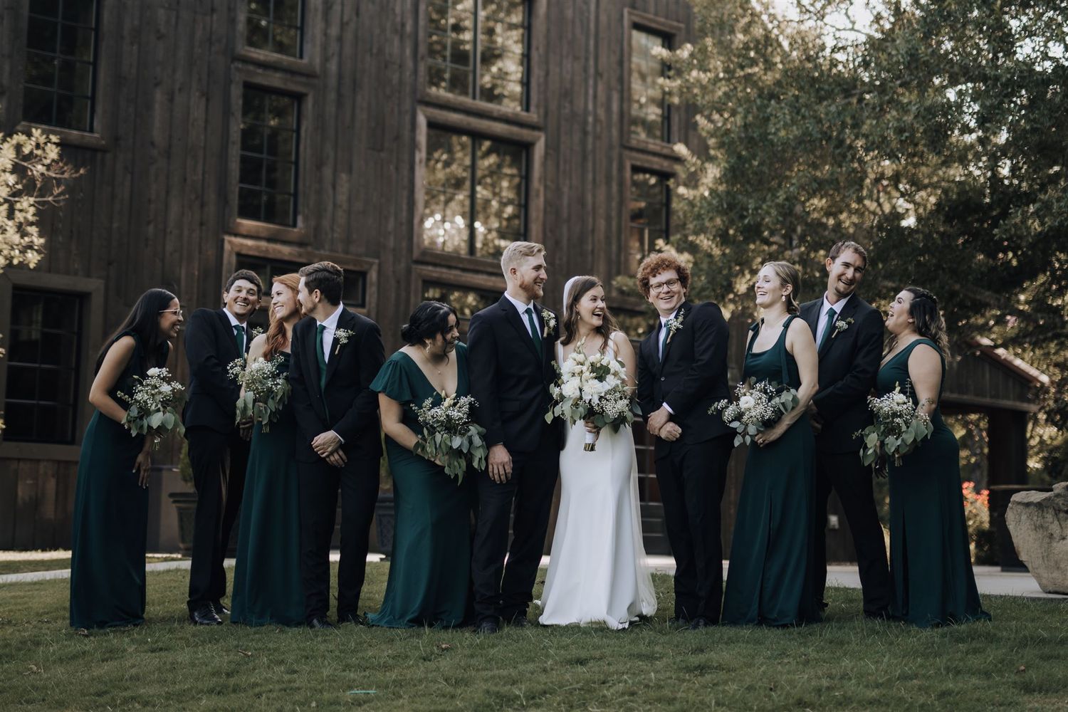 Wedding party in deep teal dresses poses with couple outside rustic barn venue.