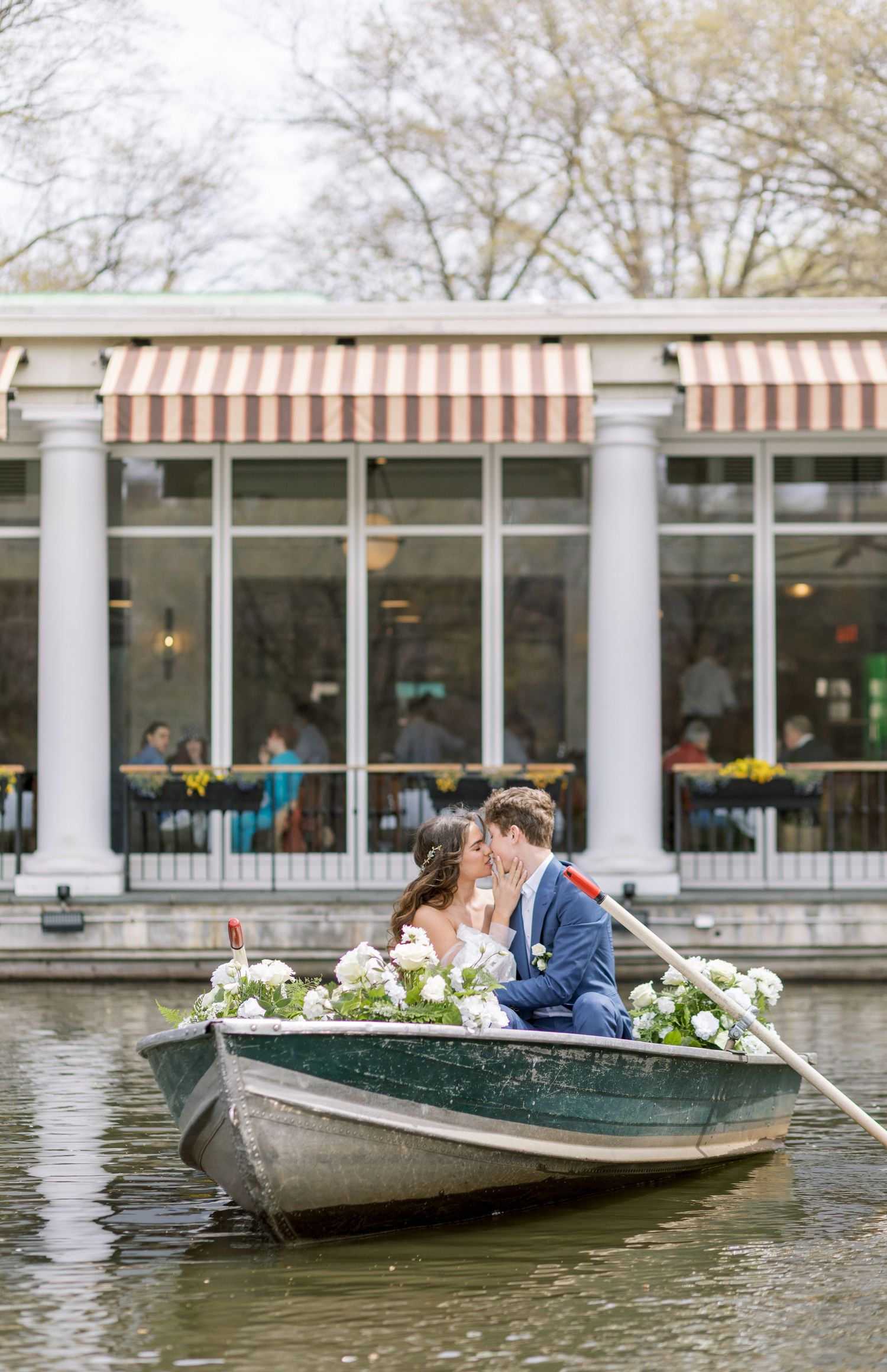 A romantic couple sits in a rowboat surrounded by spring flowers near a columned building.