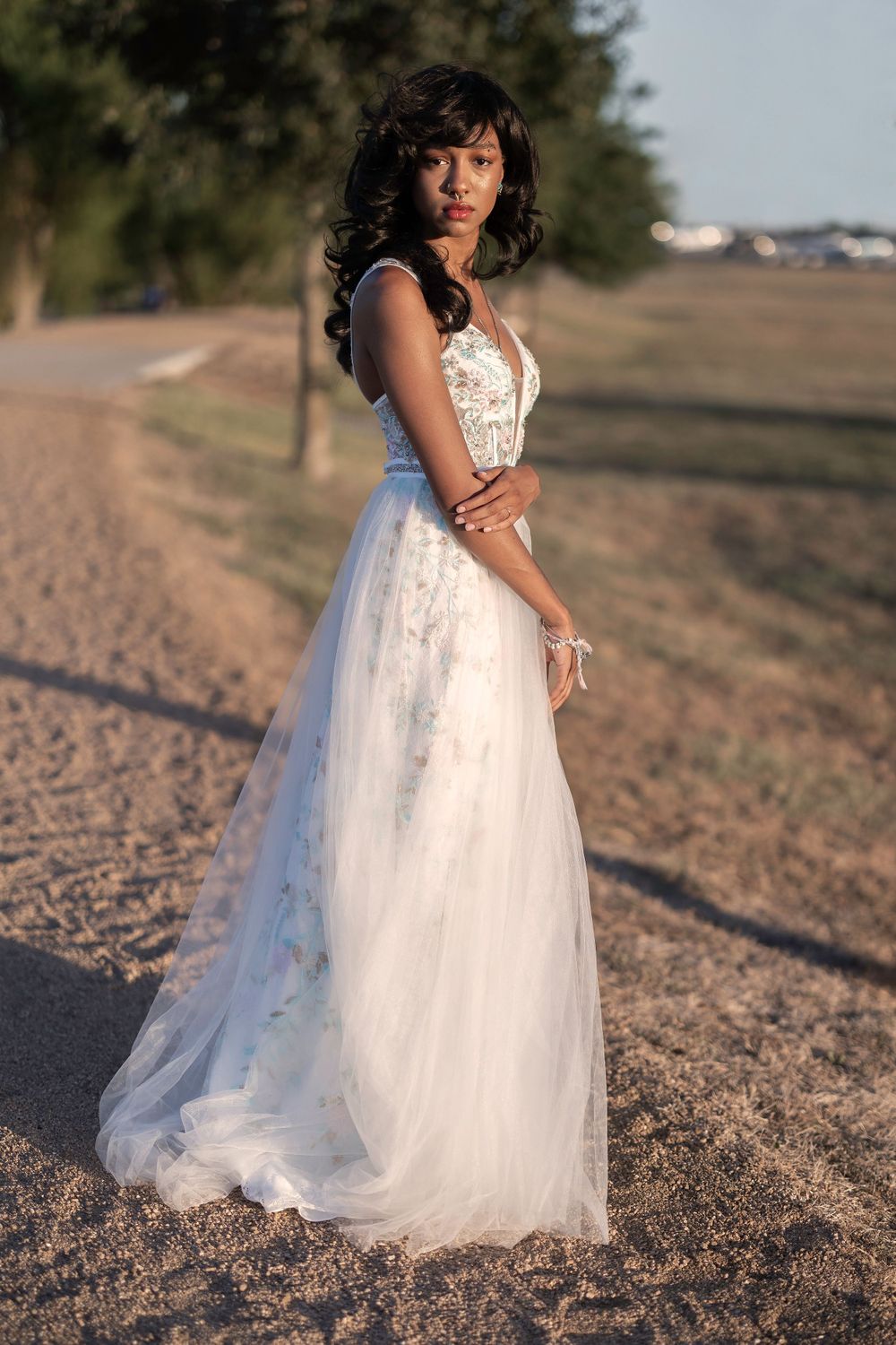 Person in flowing white wedding gown poses on dirt road during sunset.