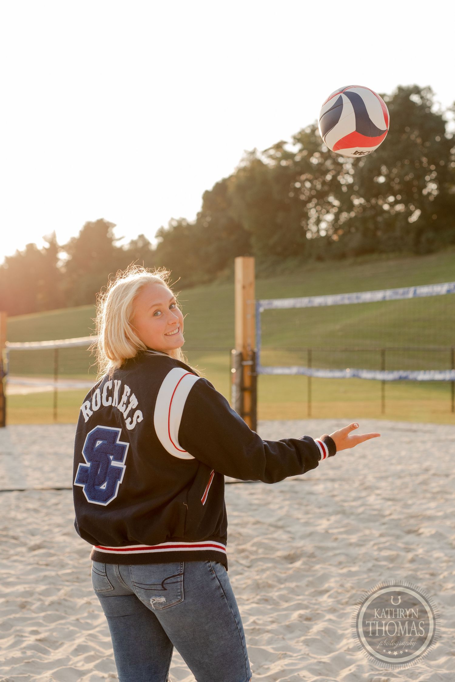 Athlete in varsity jacket playing with volleyball on sand court at sunset.