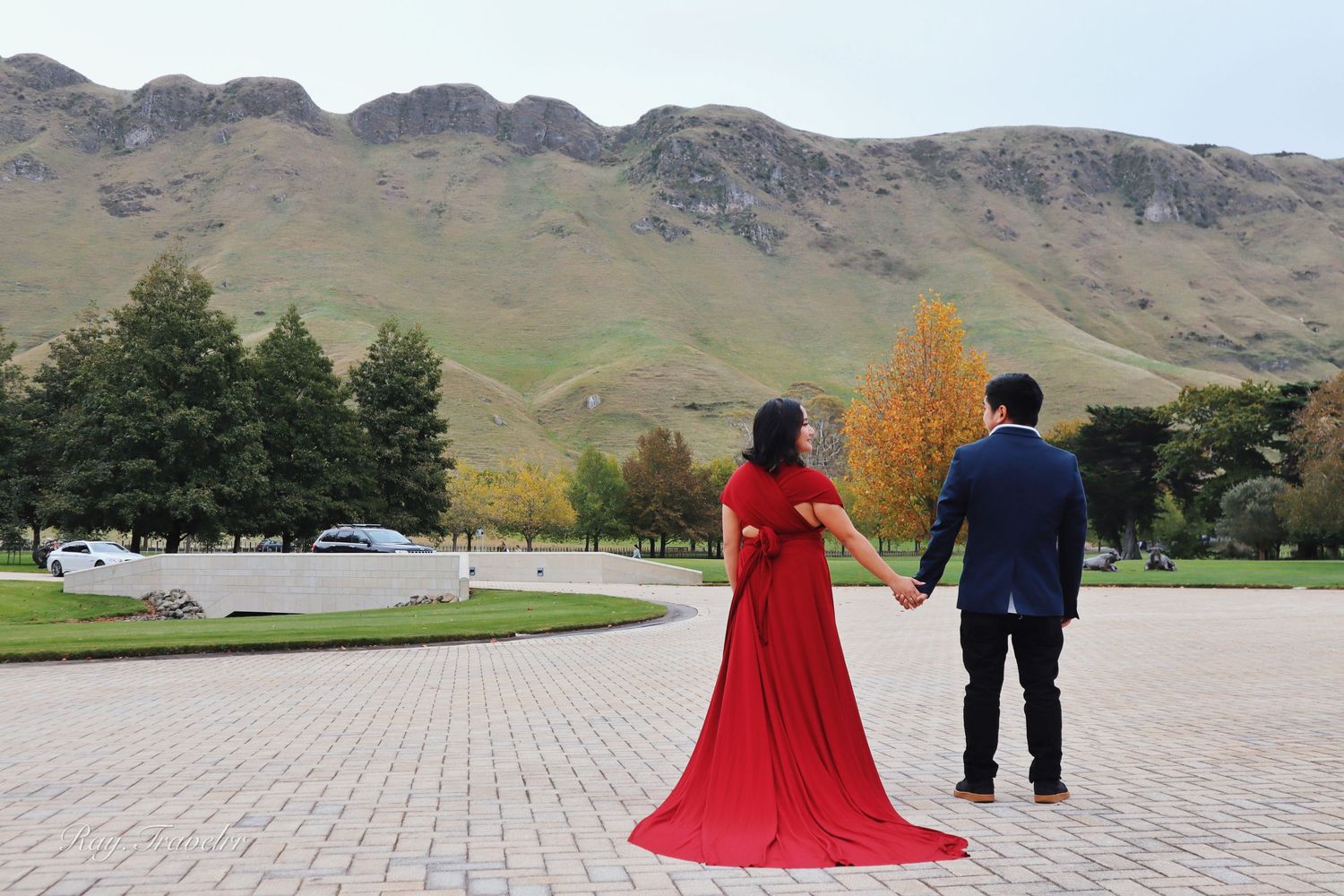 A scenic autumn mountain landscape with two figures in formal attire standing on a paved path.