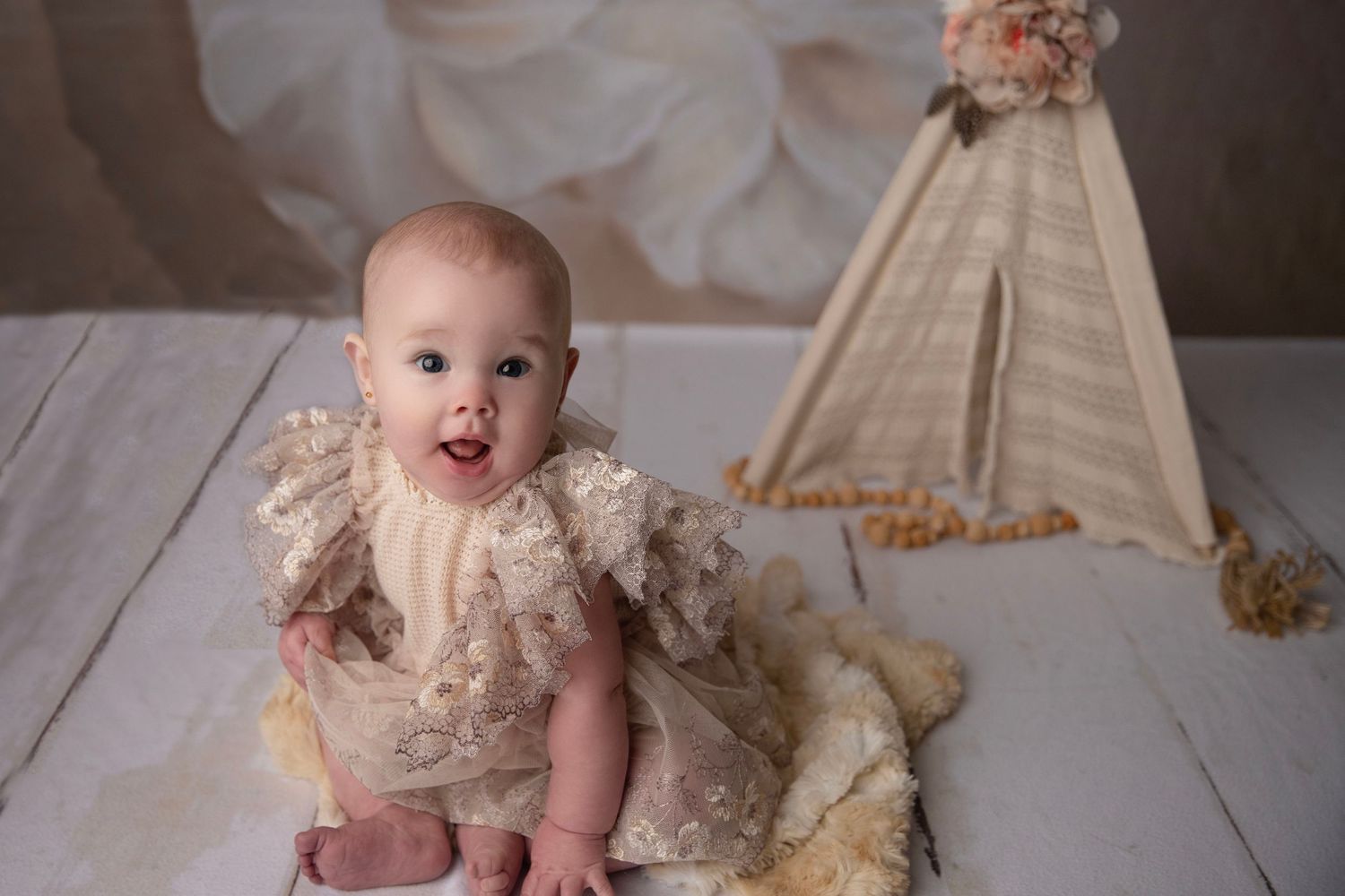 Adorable baby girl in a lace romper sitting up smiling with a teepee prop behind her.
