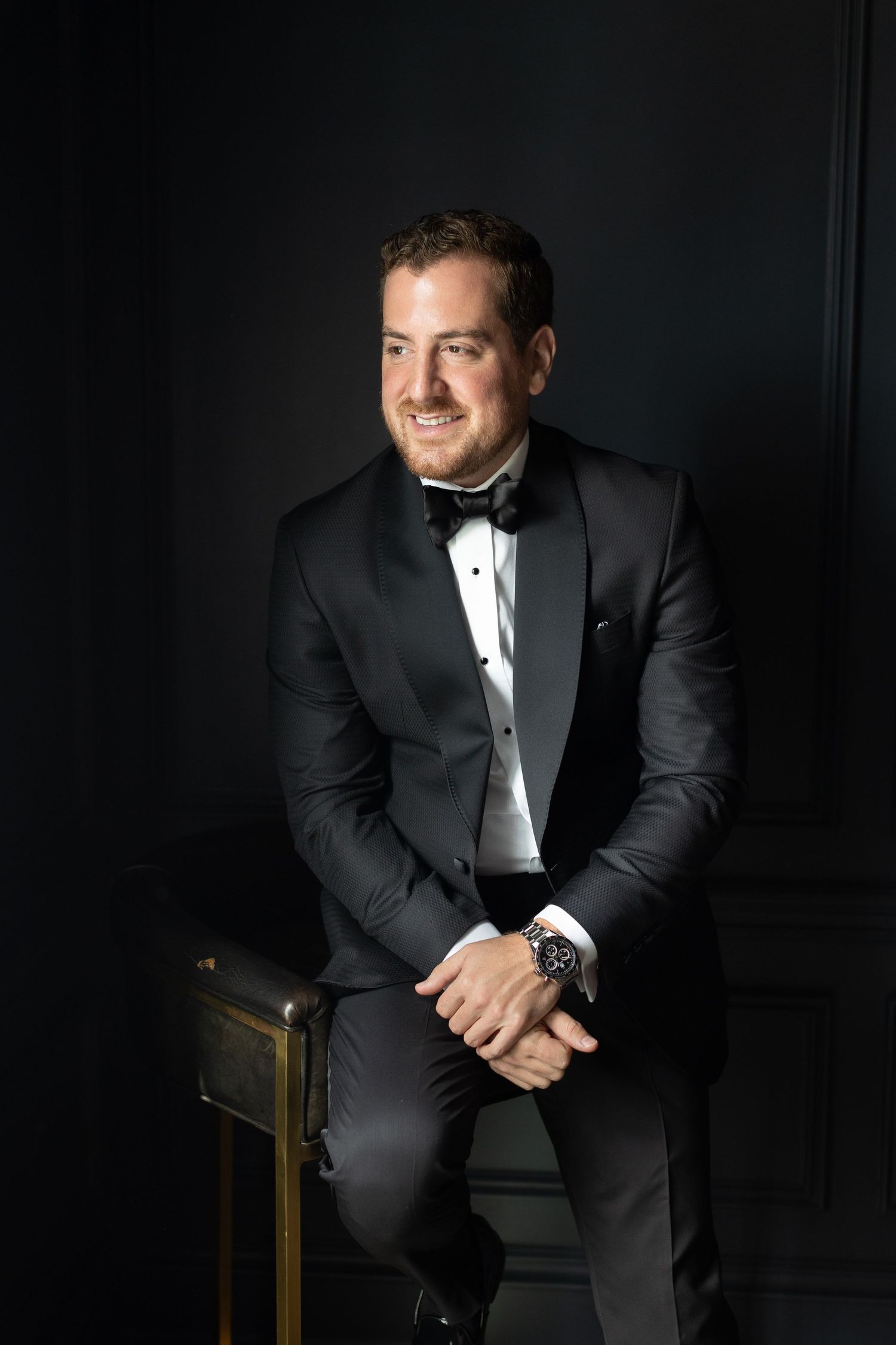 Formal portrait of groom in black tuxedo and bowtie against dark background.