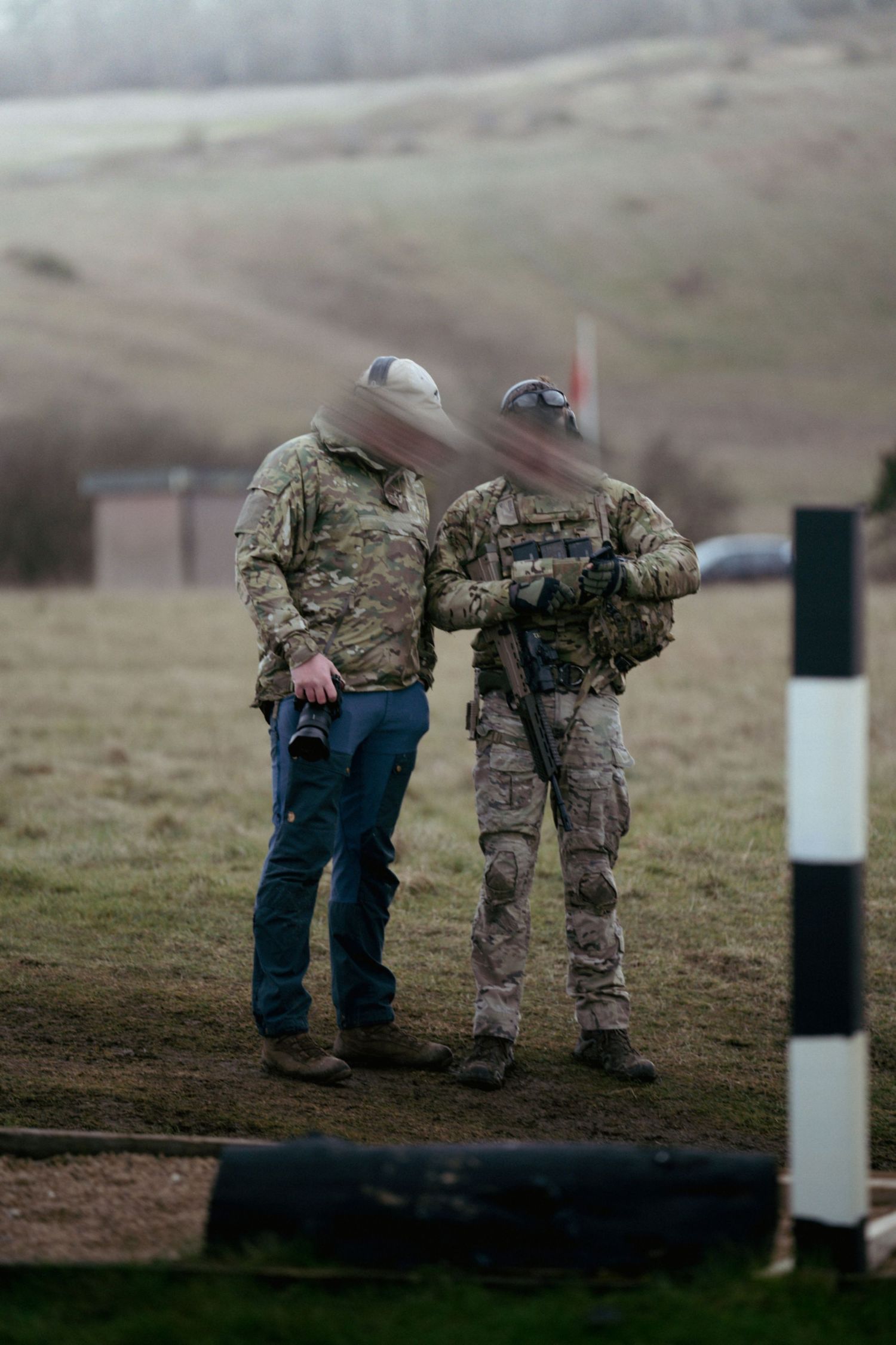 Two military personnel in tactical gear standing on grassy field near marker post.