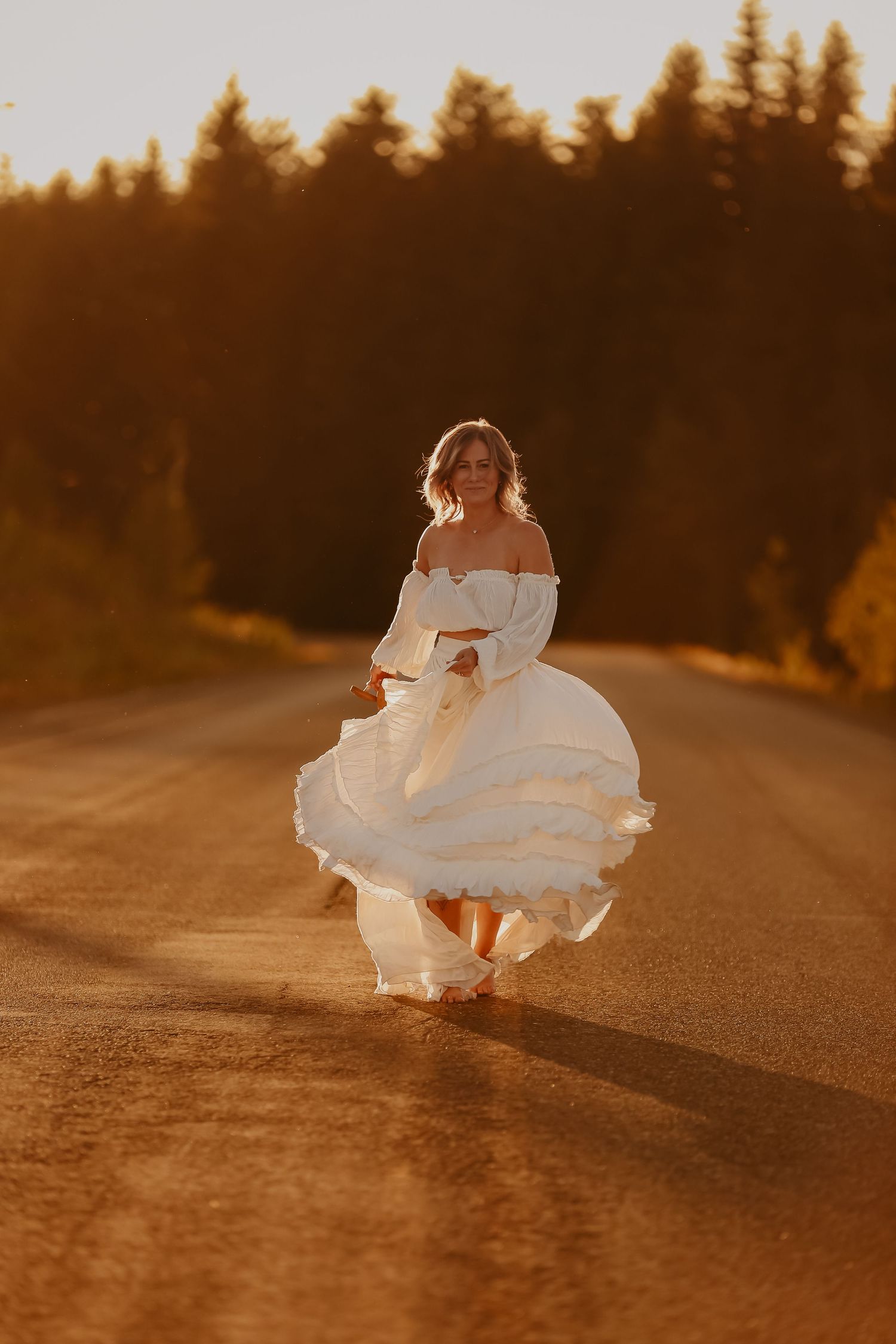 White dress flowing in golden sunset light on a rural road with evergreen trees in background.