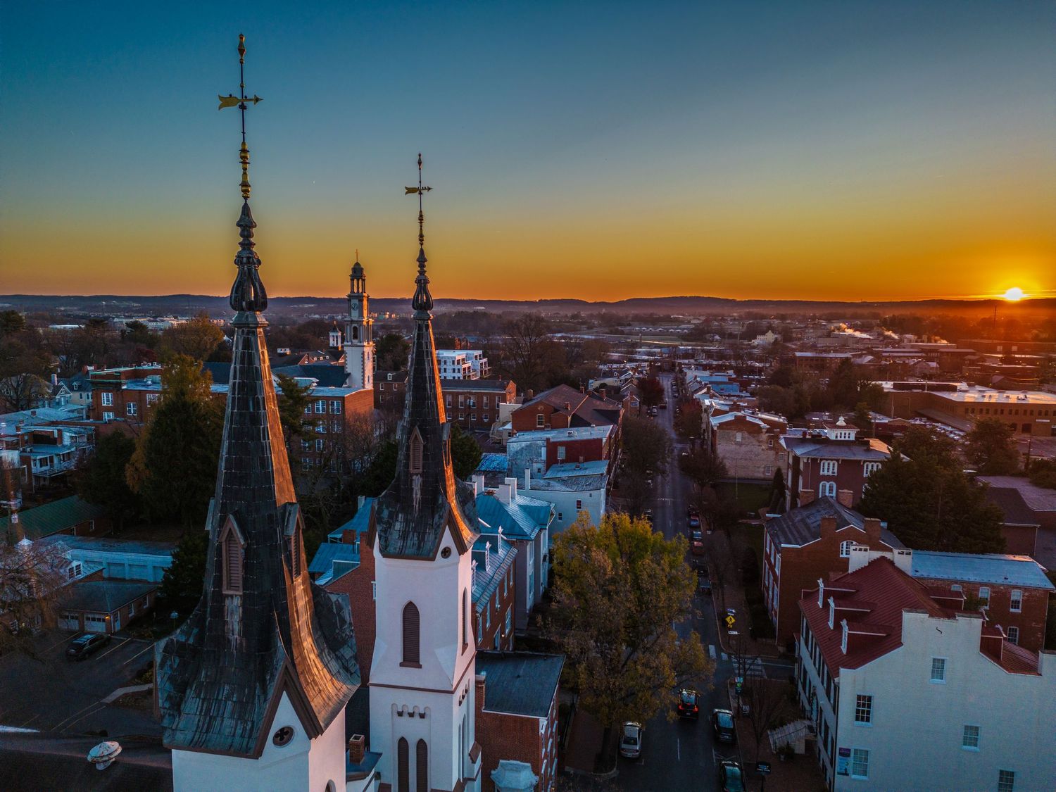Gothic church spires silhouetted against a dramatic orange sunset sky over a cityscape.