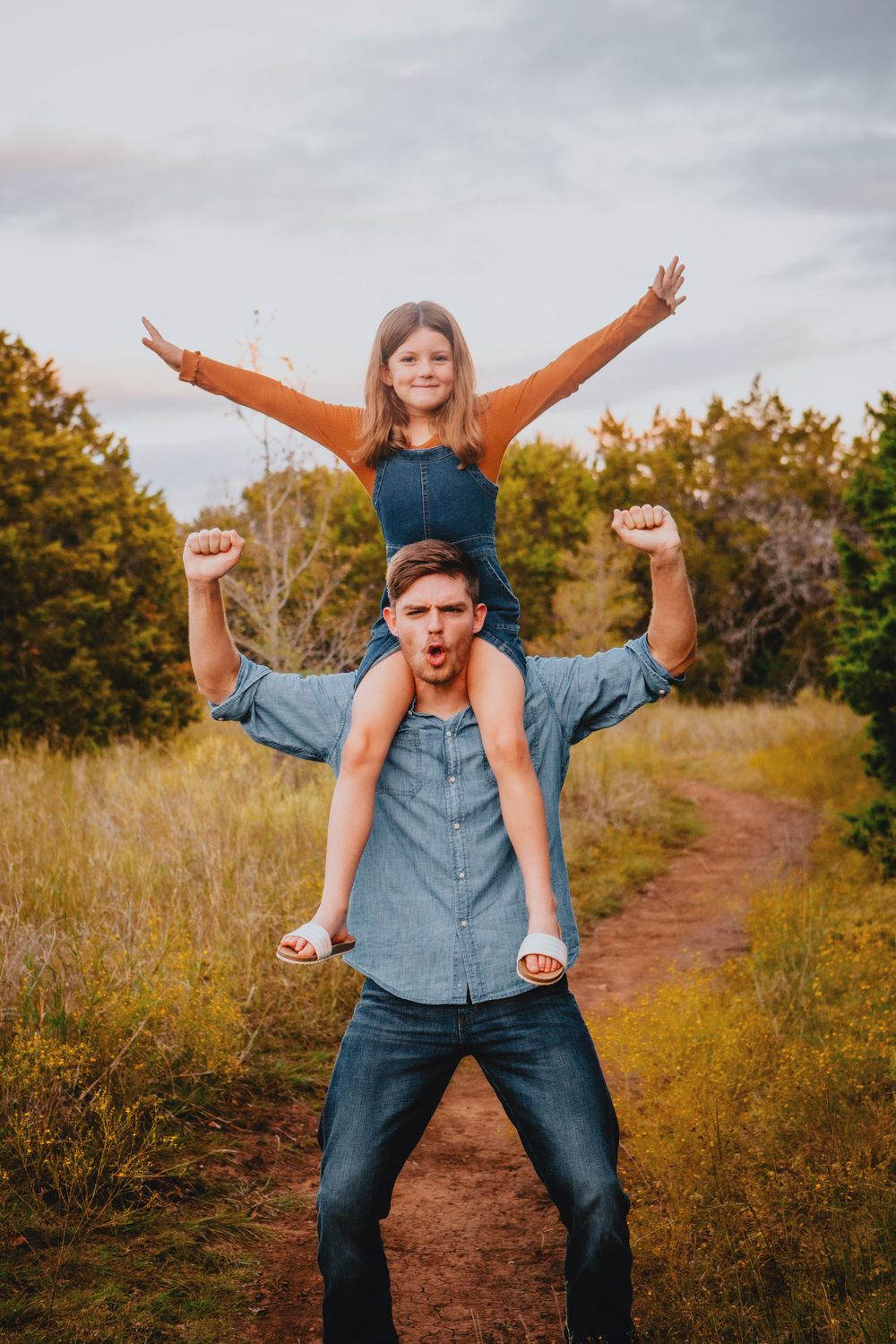 A playful outdoor scene on a dirt path with someone carrying a child in an orange shirt on their shoulders.