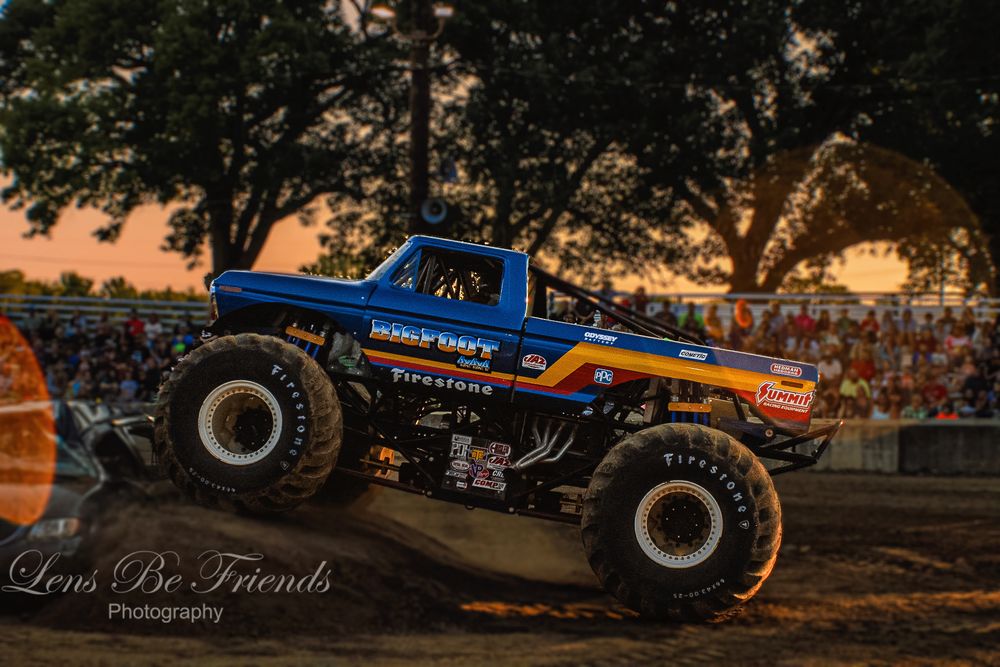 Monster truck silhouettes performing stunts at sunset with dramatic sky backdrop.