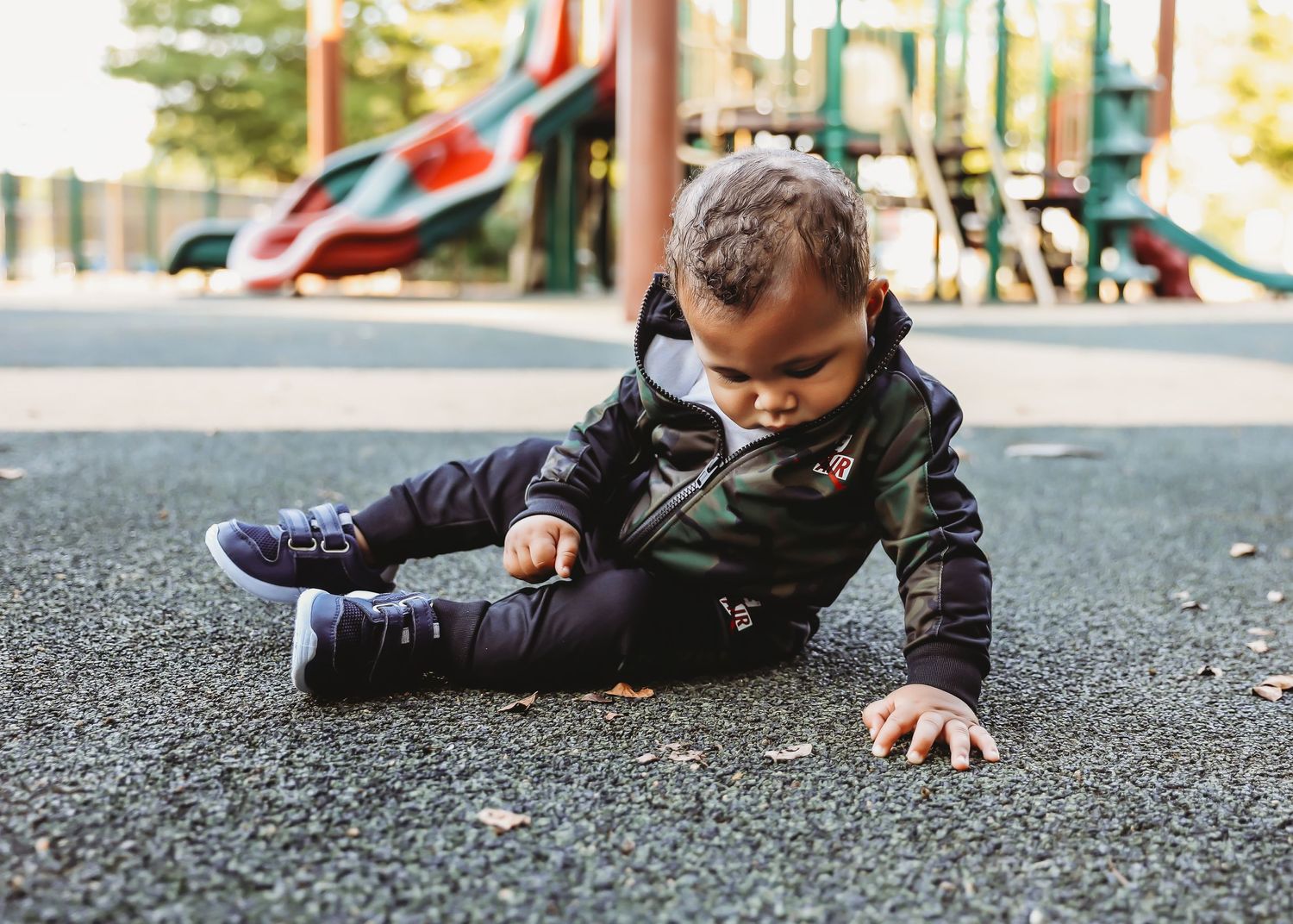 Small child wearing black jacket and purple shoes lies on playground surface near colorful equipment.