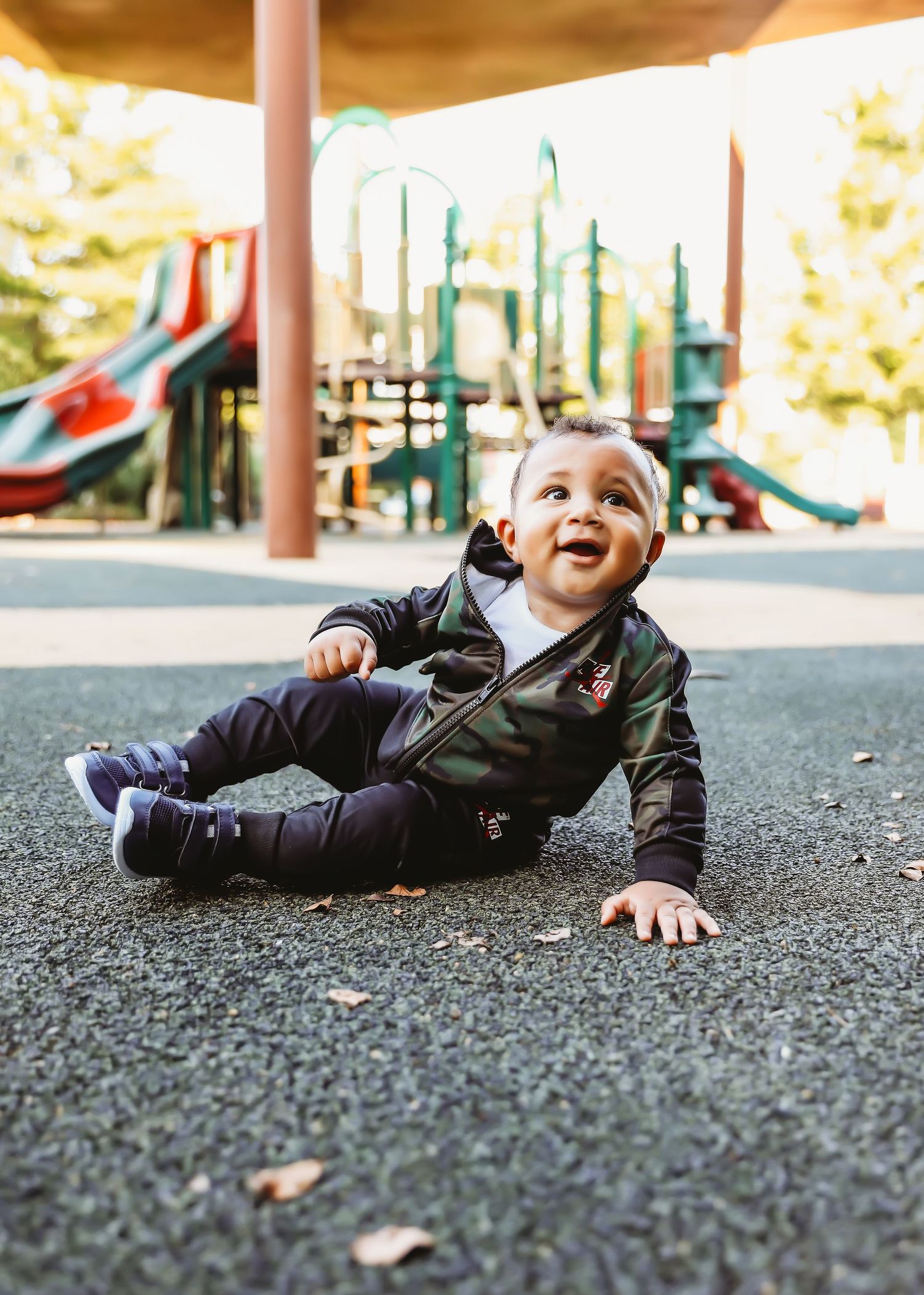 Toddler in dark jacket sits on ground beneath playground equipment with joyful expression.