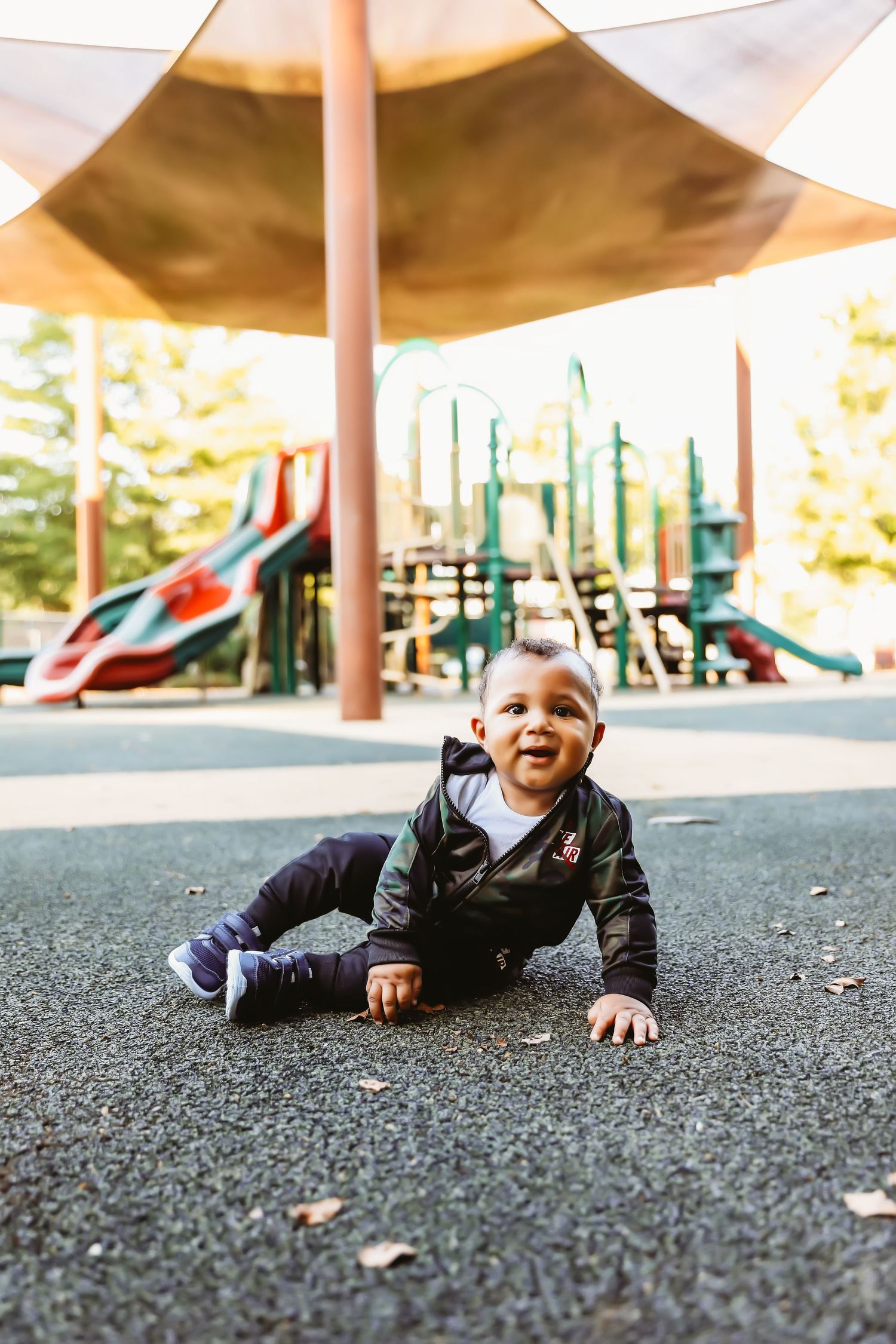 Young child in black jacket laughs while playing under shade structure at playground.