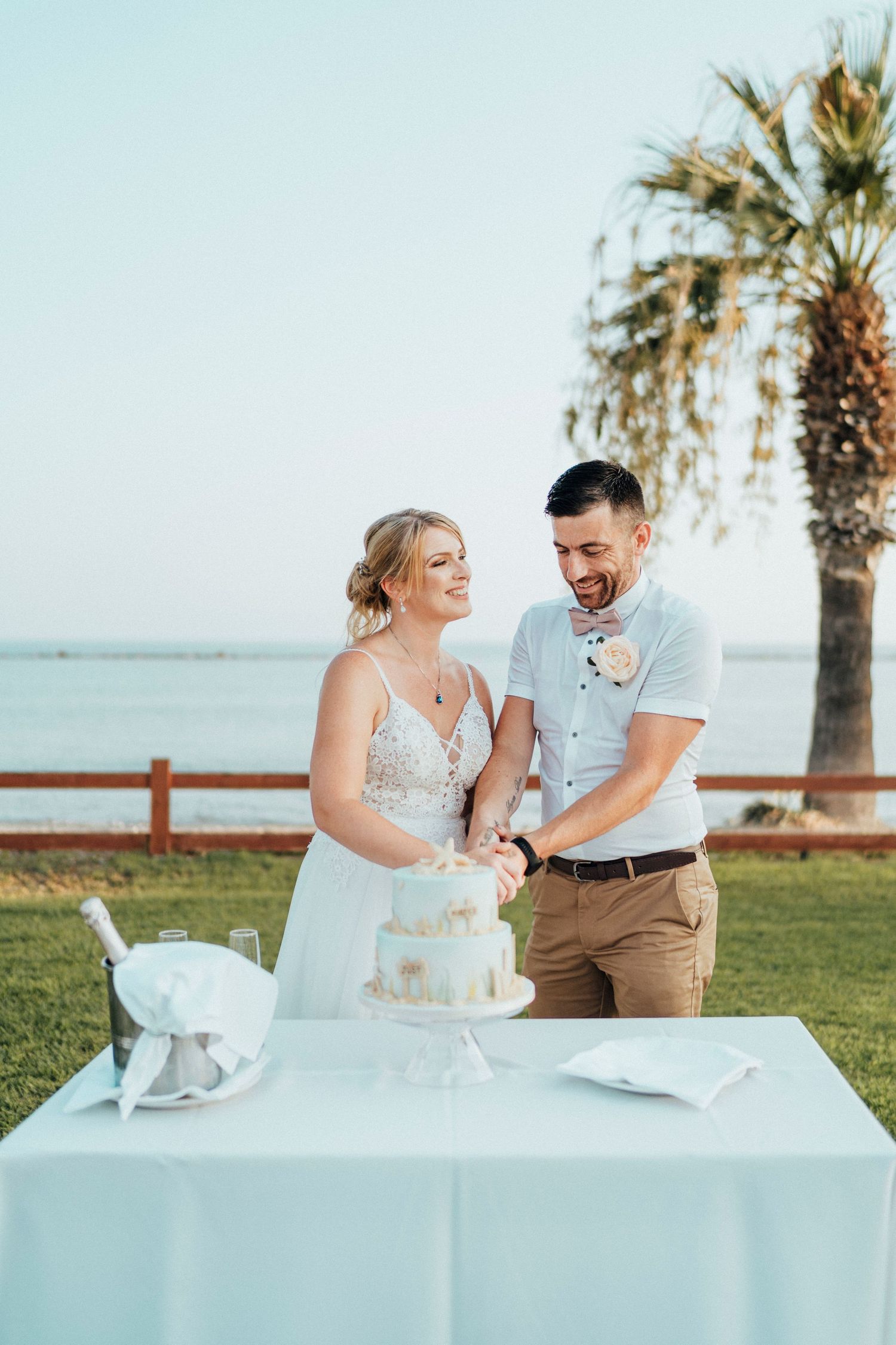 A couple cuts their elegant white wedding cake together at an outdoor reception by the ocean with palm trees.