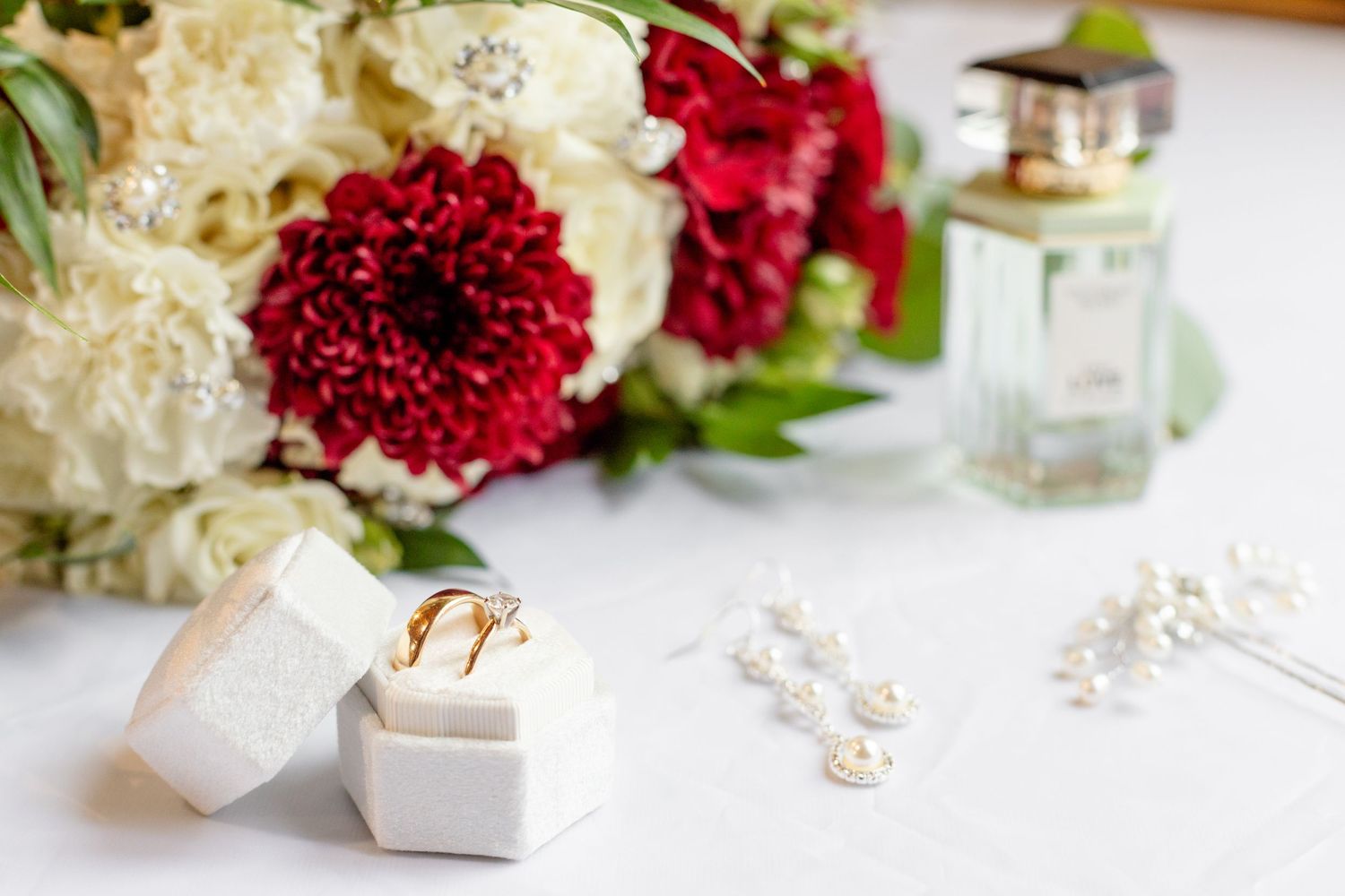 Wedding details with red and white flowers, rings, and jewelry on white surface.