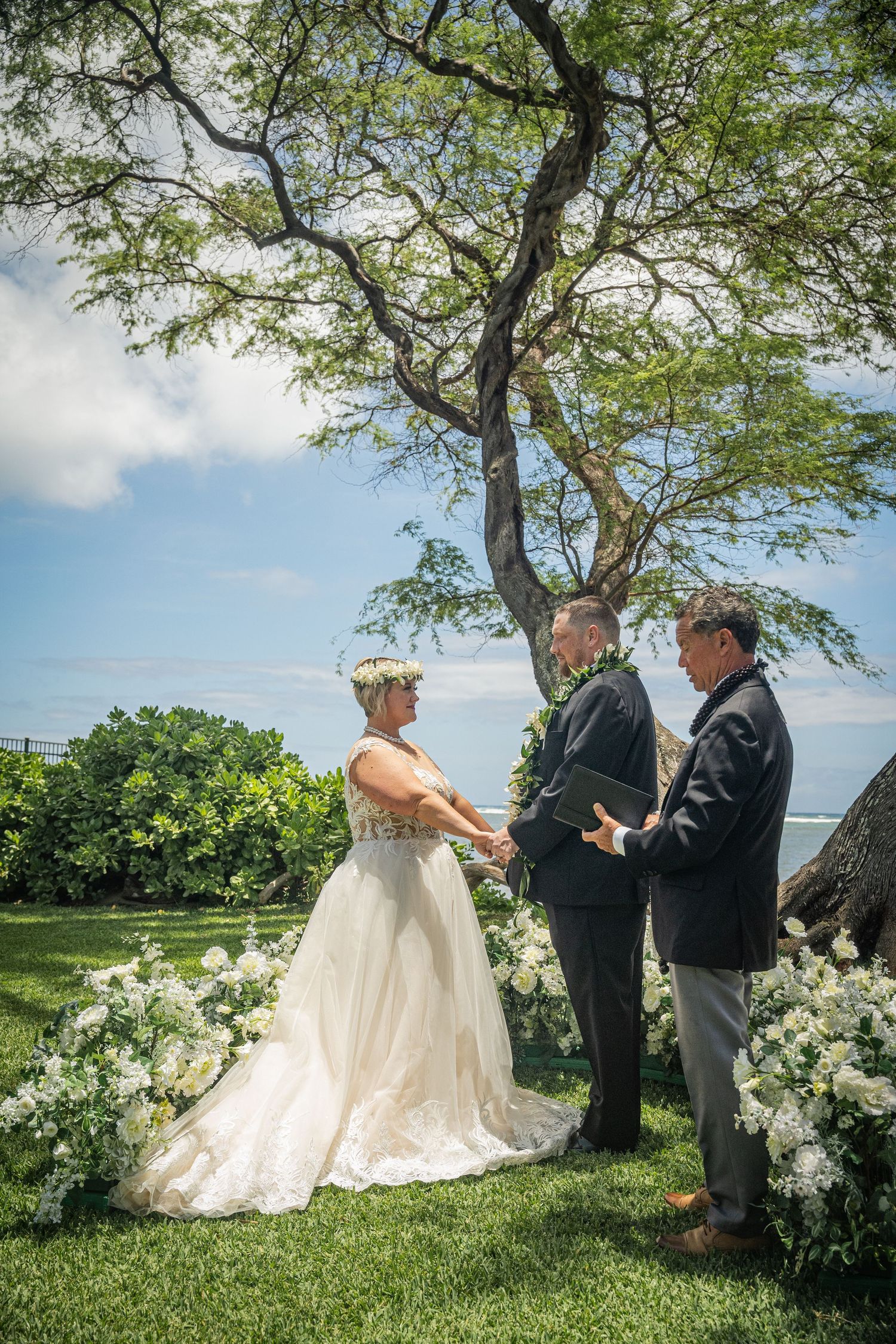 Outdoor wedding ceremony under large tree with white floral arrangements and ocean backdrop.