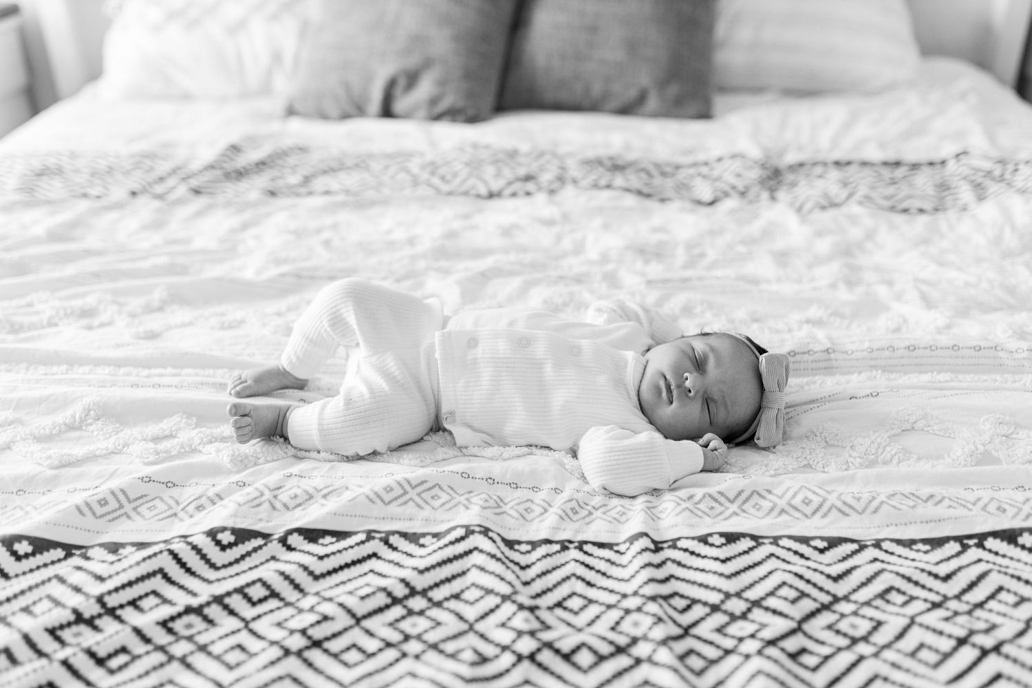Black and white portrait of a newborn baby in white clothing resting on patterned bedding.
