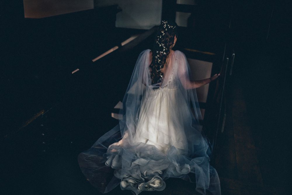 Moody portrait of a bride in a flowing dress seated in dark atmospheric lighting.