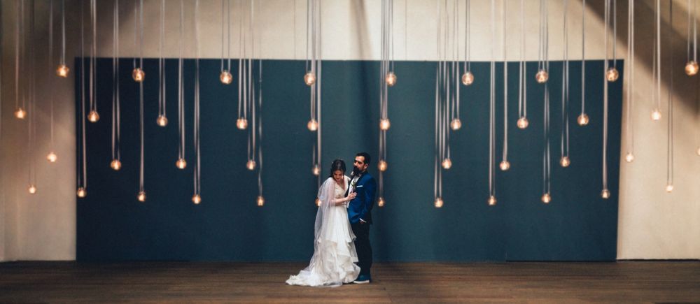Wedding couple posing against a wall decorated with hanging string lights.