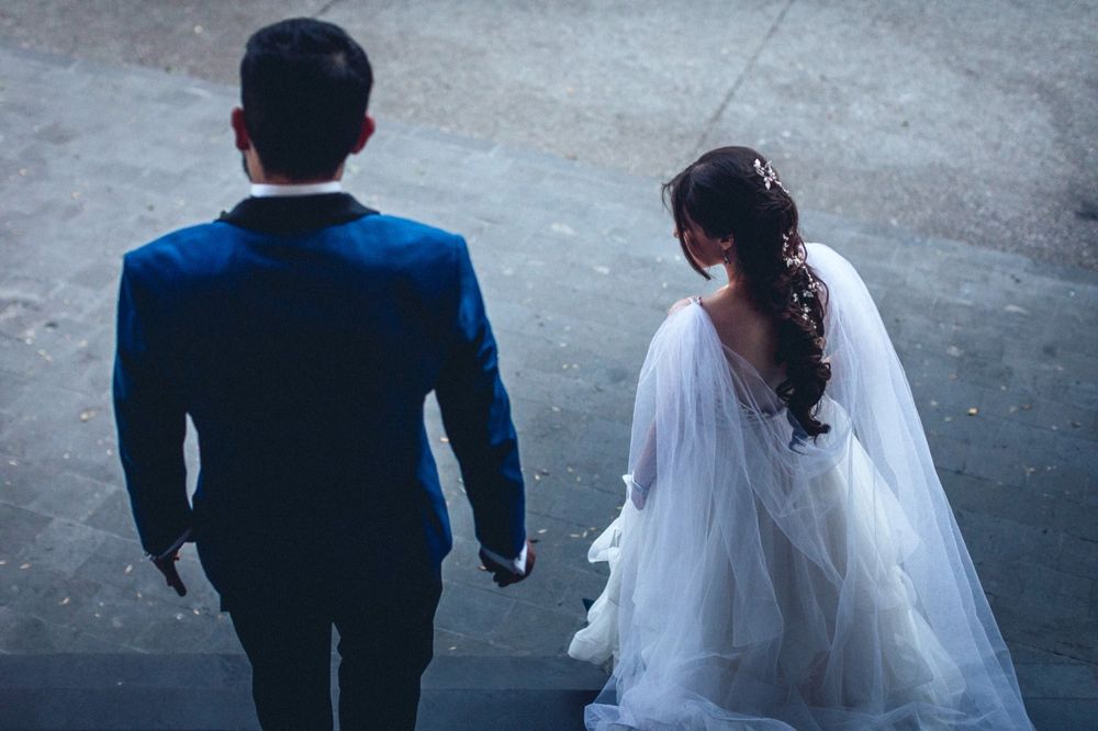 A bride in a flowing veil and groom in navy suit walking together viewed from behind.