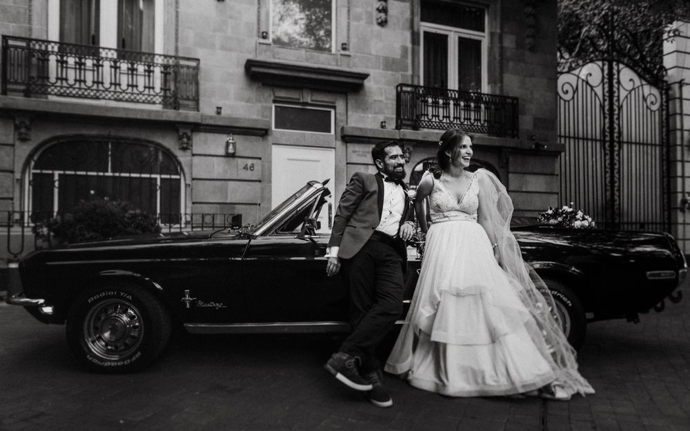Stylish wedding couple posing beside a classic vintage car on a city street.