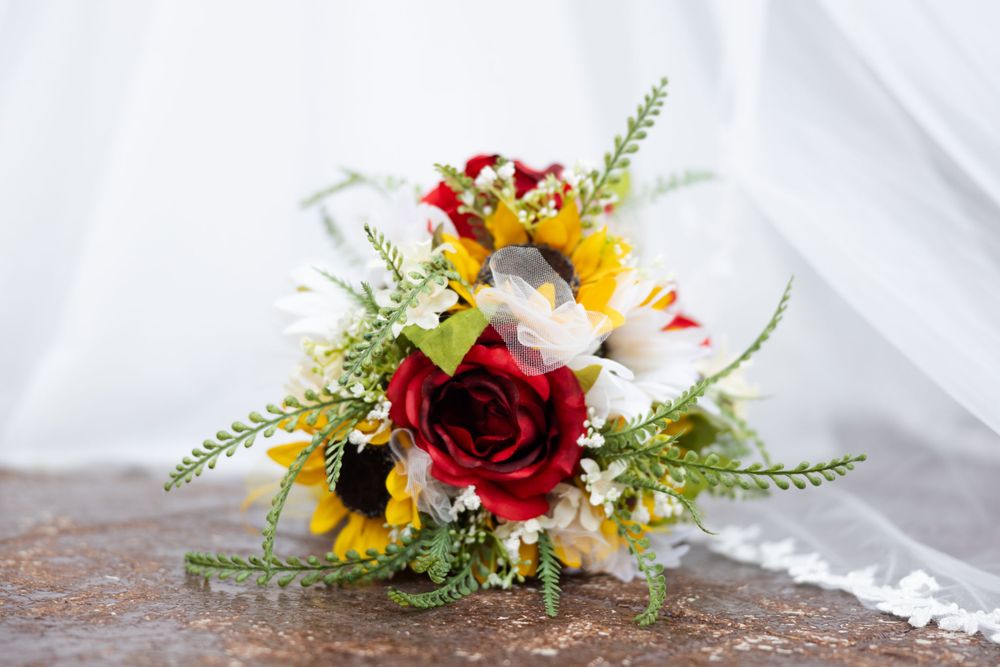 A vibrant wedding bouquet with red roses, sunflowers, and white wildflowers on a rustic surface.