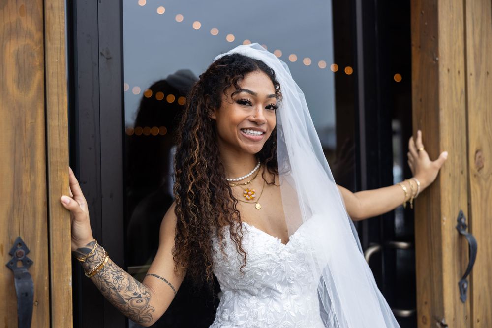 A bride stands between wooden doors with string lights visible in the background.