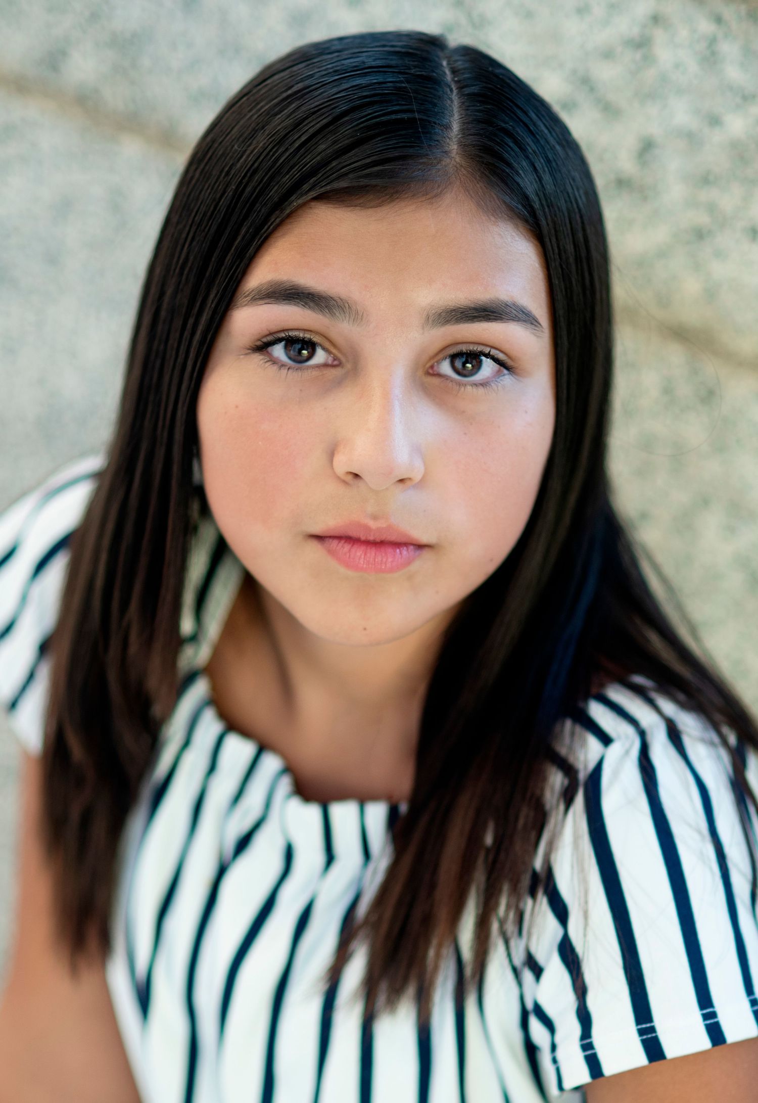 Young person in striped shirt looking at camera with natural makeup and long dark hair against stone background.