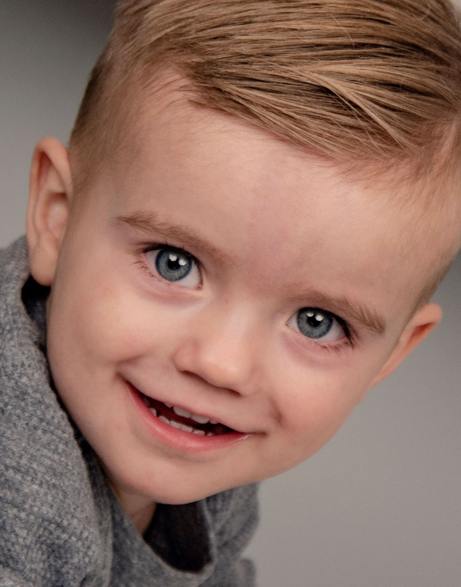 A child with bright blue eyes and light brown hair wearing a gray sweater smiles warmly at the camera.