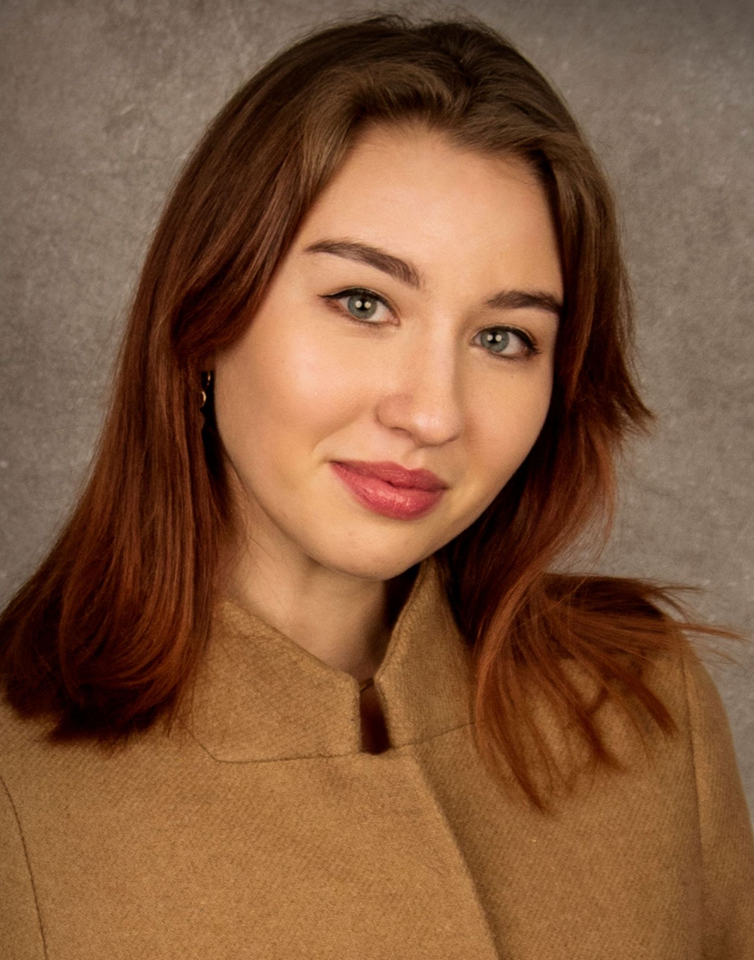 Portrait shot showing a smiling woman wearing a brown sweater against a gray backdrop.