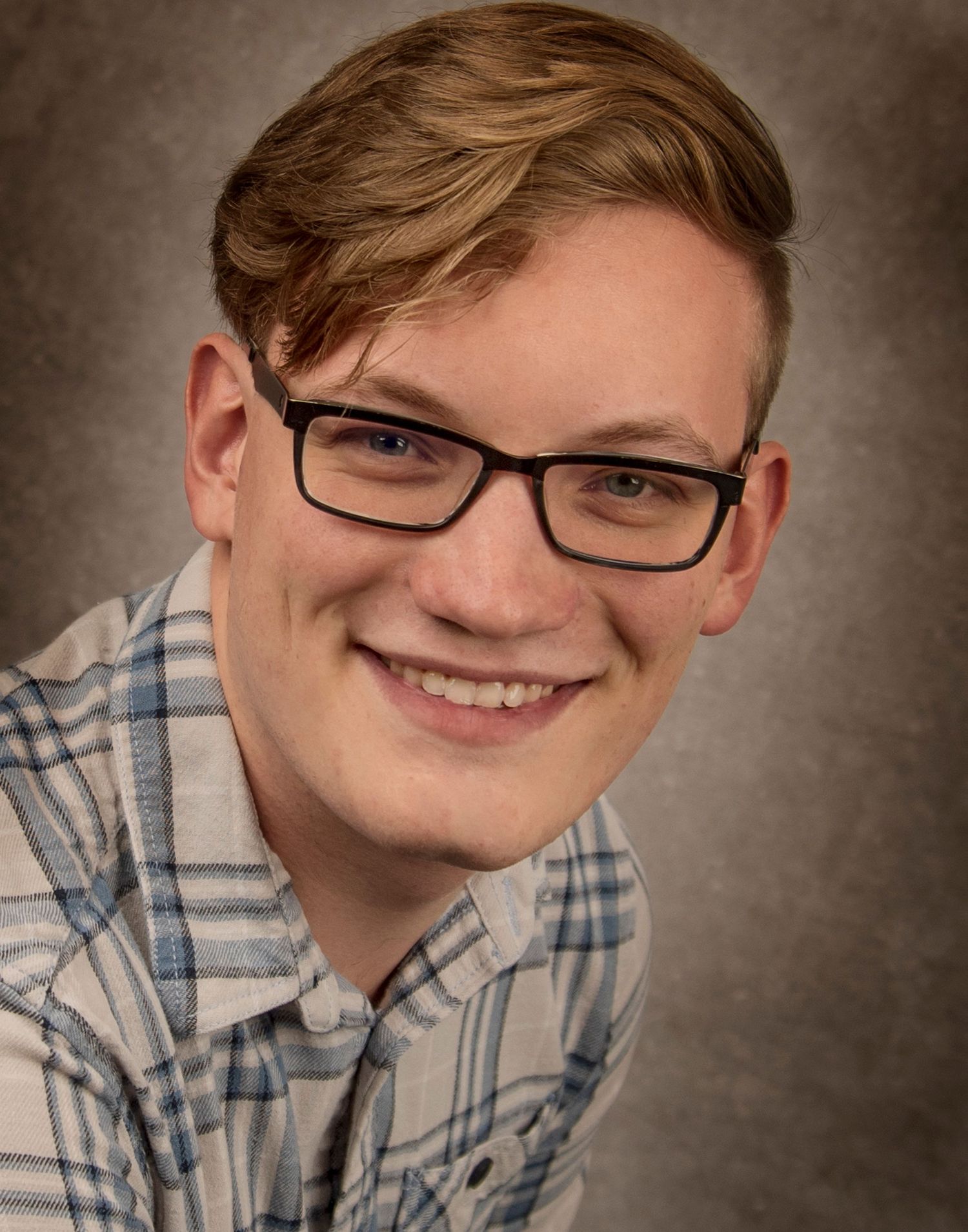 Professional headshot of a person smiling warmly wearing glasses and a plaid button-down shirt against a brown backdrop.