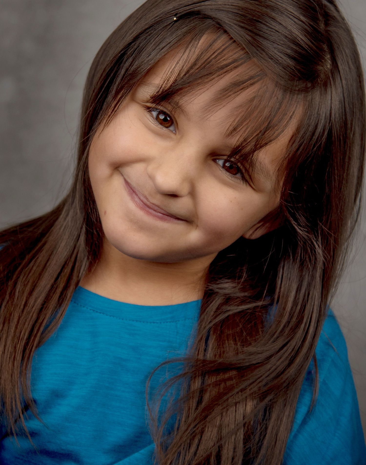 A portrait of a young child wearing a bright blue shirt with long dark hair and bangs smiling warmly at the camera.