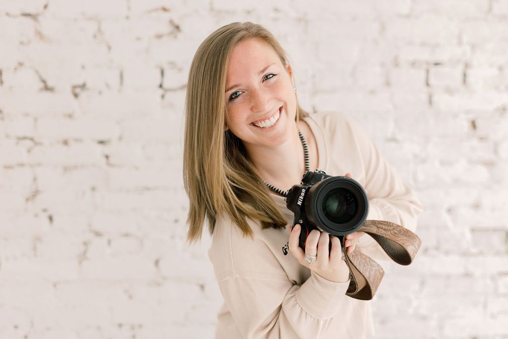 Professional photographer holding a camera while wearing a cream colored sweater against a white brick wall.