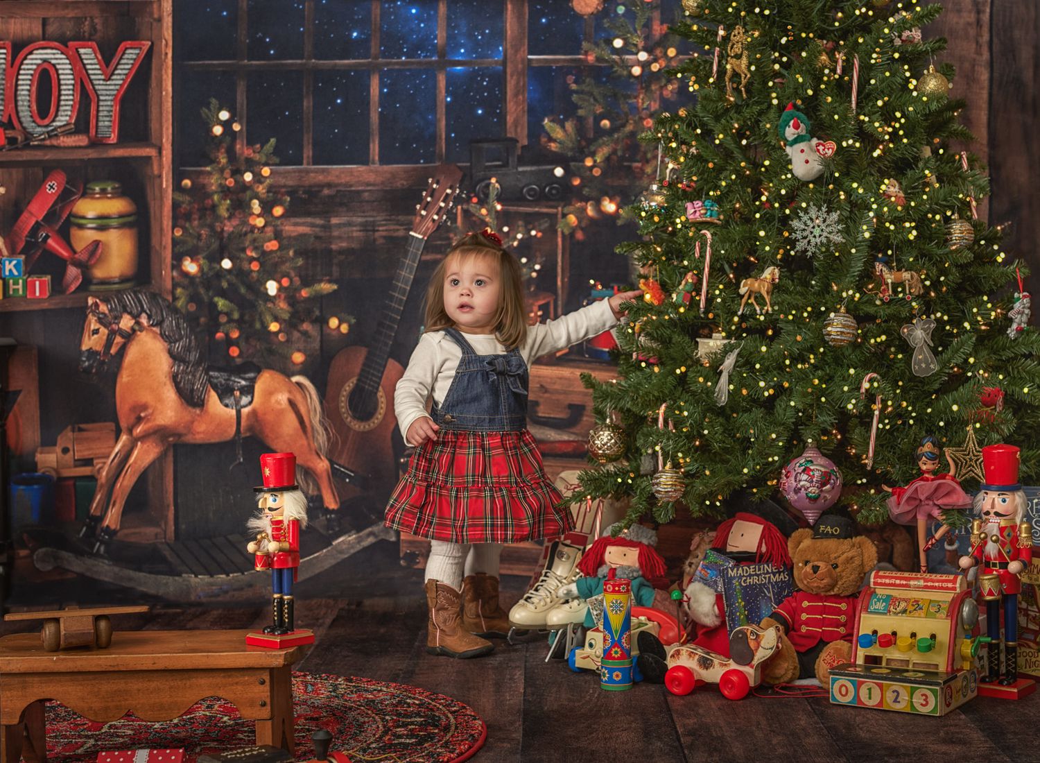 Toddler girl in country jumper and cowgirl boots adding an ornament to Christmas tree that is surrounded by toys