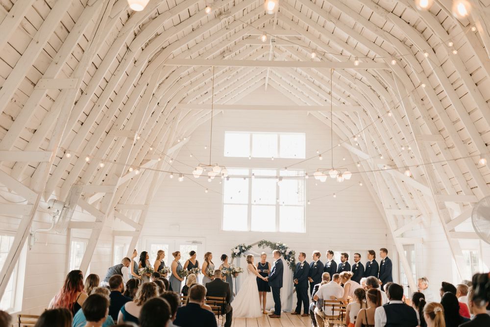 Wide shot of wedding ceremony in stunning white barn venue with string lights and full wedding party.