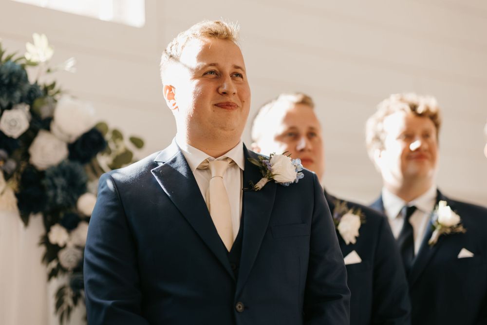 Emotional groom watches his bride walk down the aisle wearing a navy suit with white boutonniere.