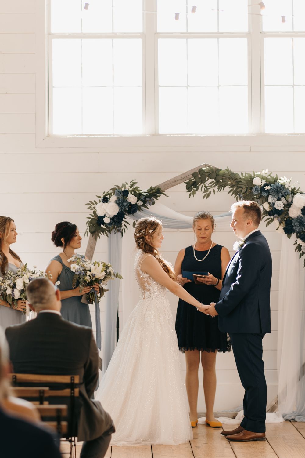 Bride and groom hold hands during indoor wedding ceremony beneath a greenery and white floral arch.