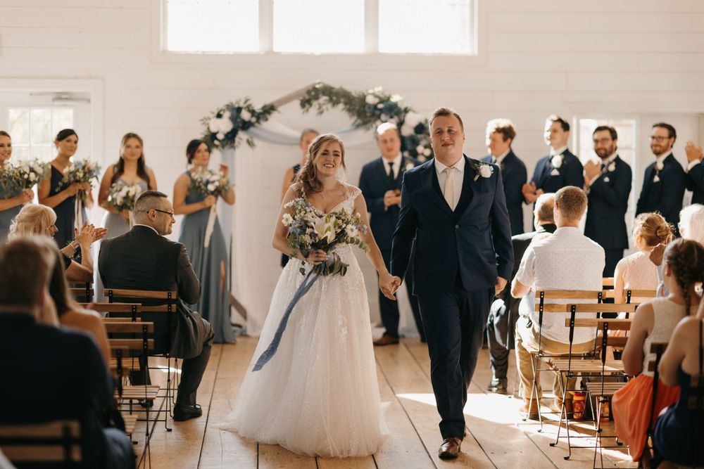 Bride and groom walk hand in hand down the aisle after wedding ceremony in white barn venue.