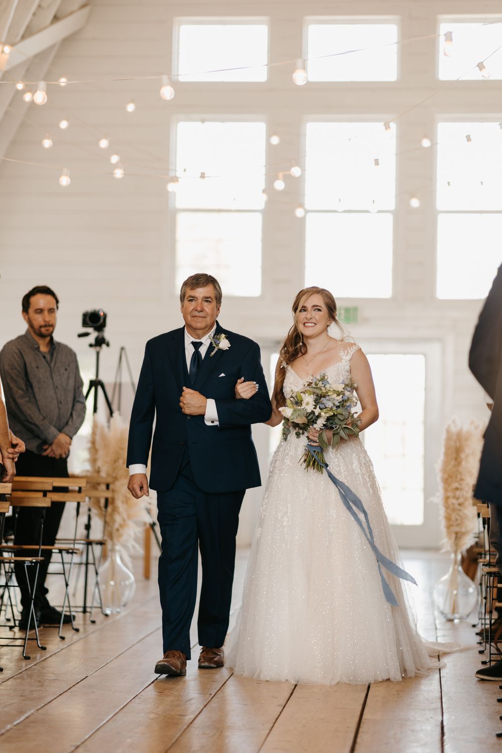 Bride and father walk down the aisle past seated guests in a sunlit white barn wedding ceremony.