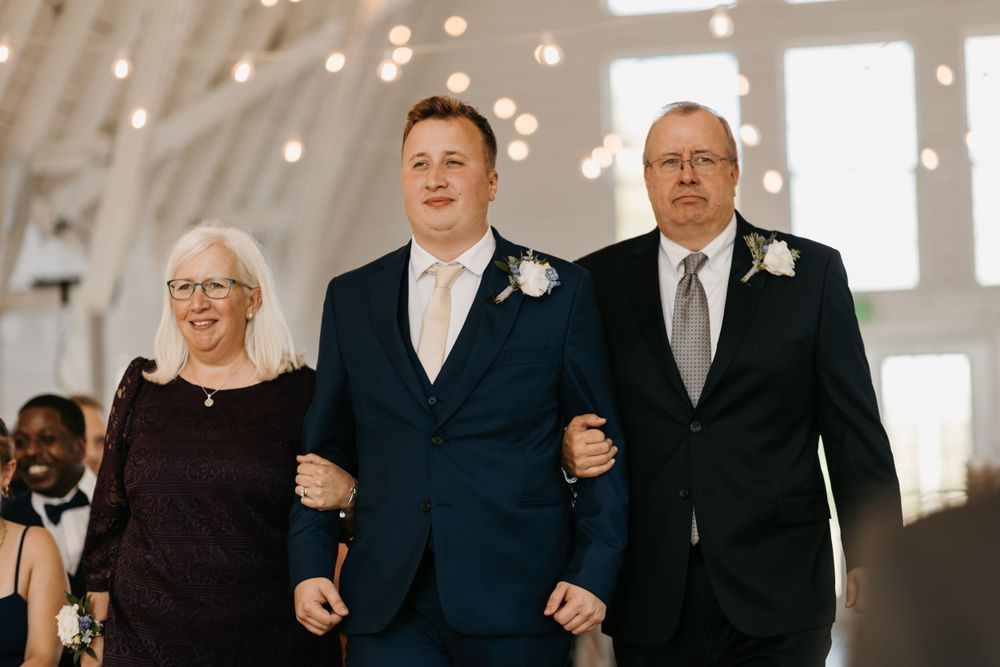Groom in navy suit walks proudly with his parents down the aisle at an elegant wedding ceremony.
