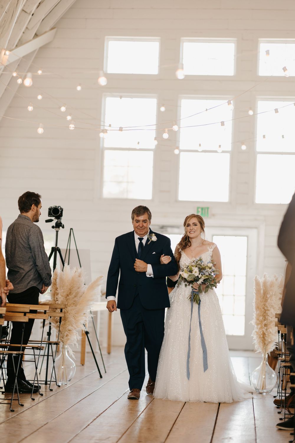 Father escorts bride down the aisle in elegant white barn venue with string lights and pampas grass decor.