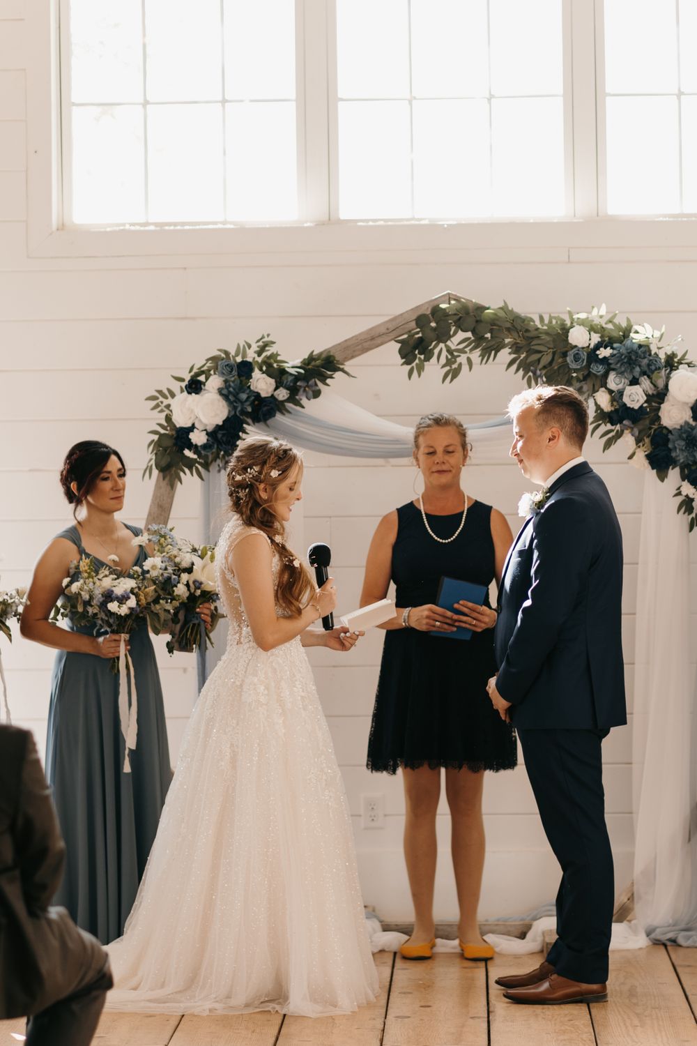 Bride reads vows to groom at indoor wedding ceremony beneath floral arch with white and blue flowers.