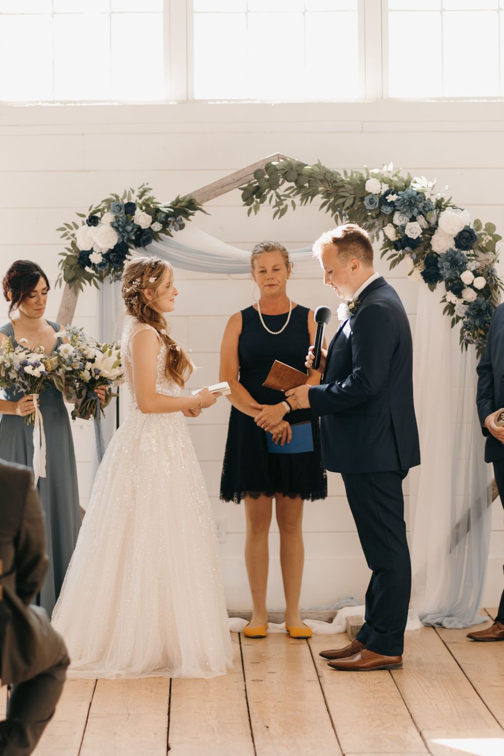 Bride and groom hold hands at floral arch altar as officiant reads vows in bright white barn venue.