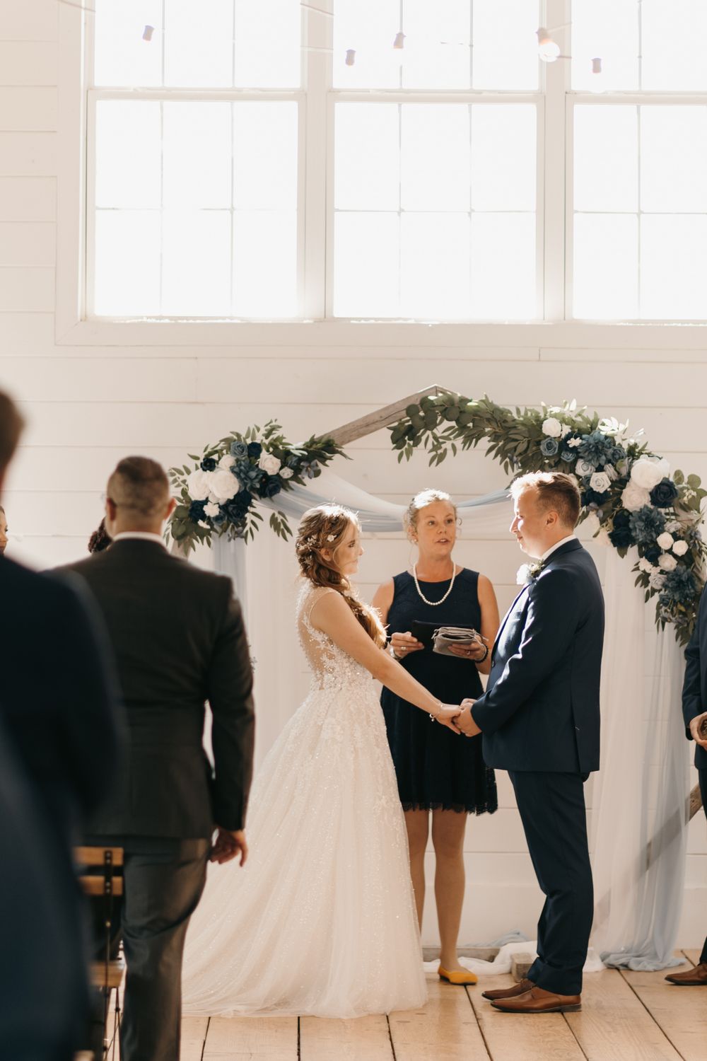 Bride and groom exchange vows at floral arch altar in white barn venue with female officiant.