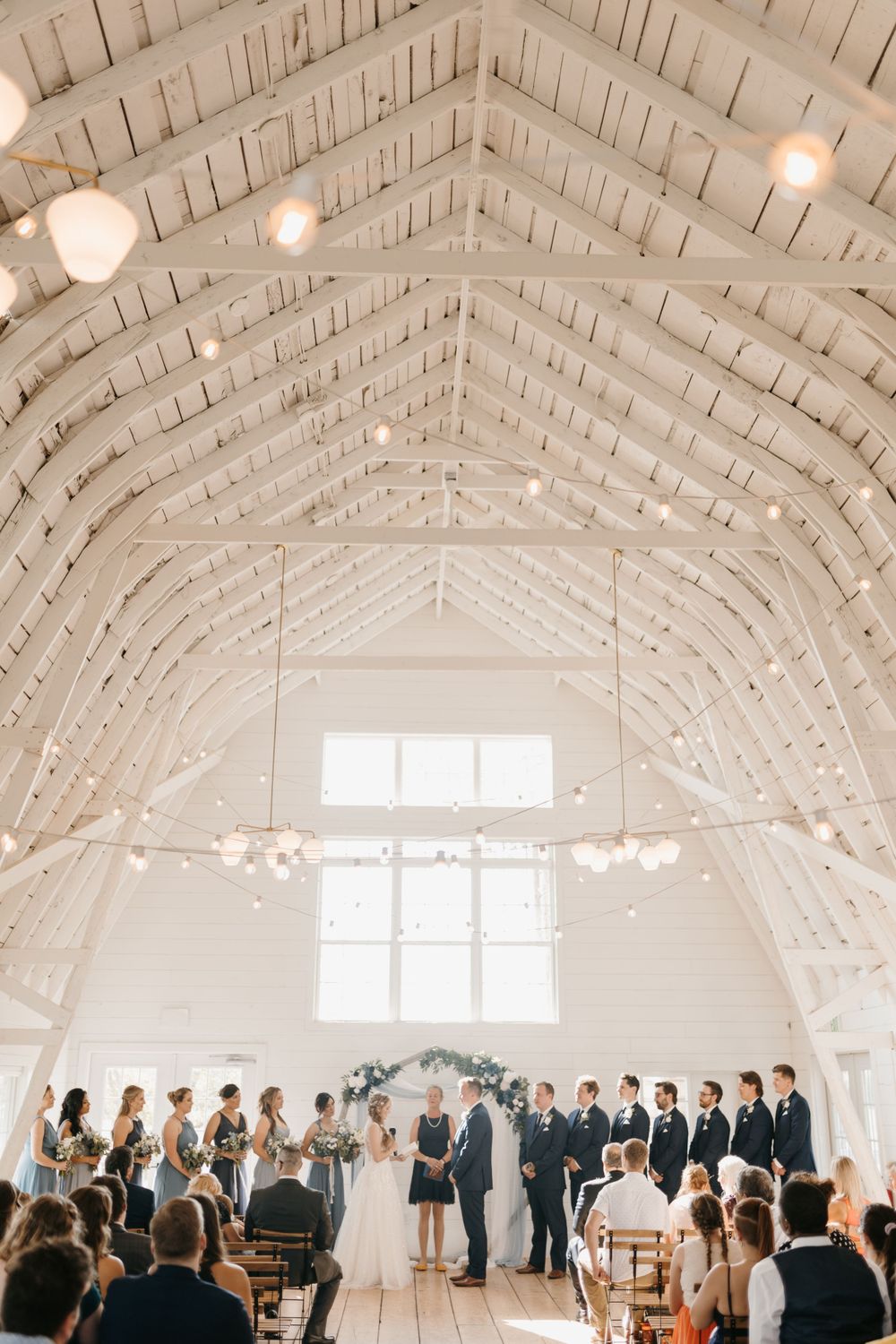 Panoramic view of elegant white barn wedding ceremony with vaulted ceiling and string lights.